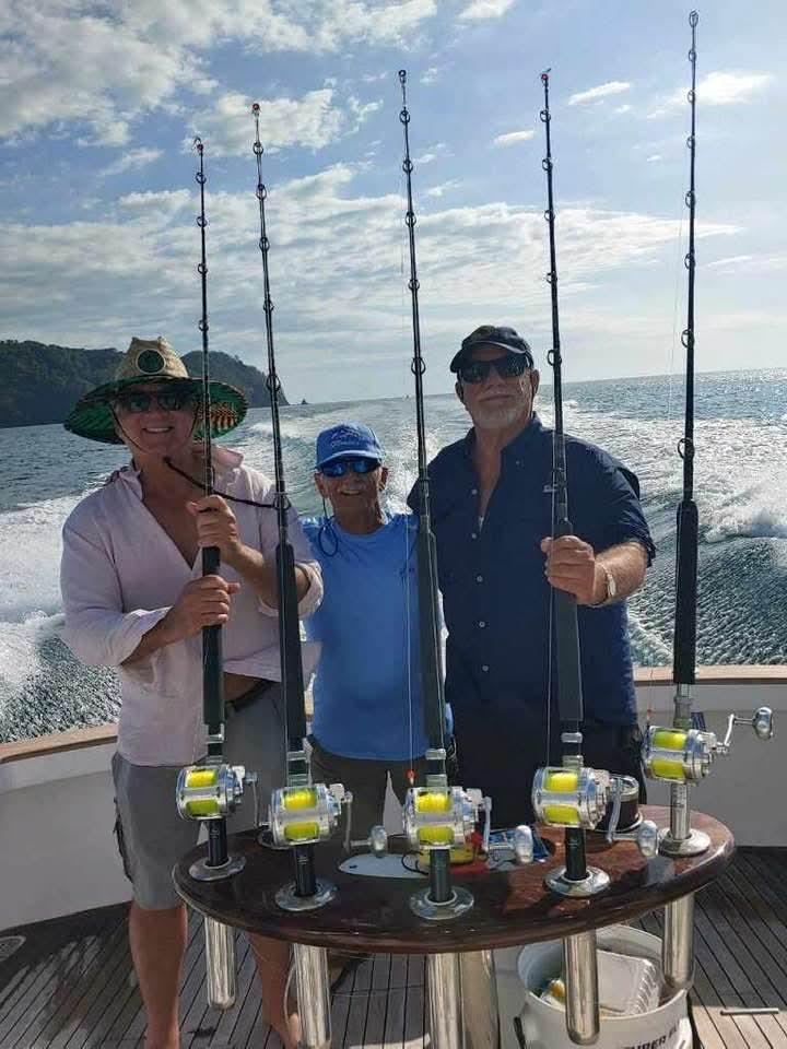 Three men hold fishing rods on a boat while the sun shines brightly and waves splash.