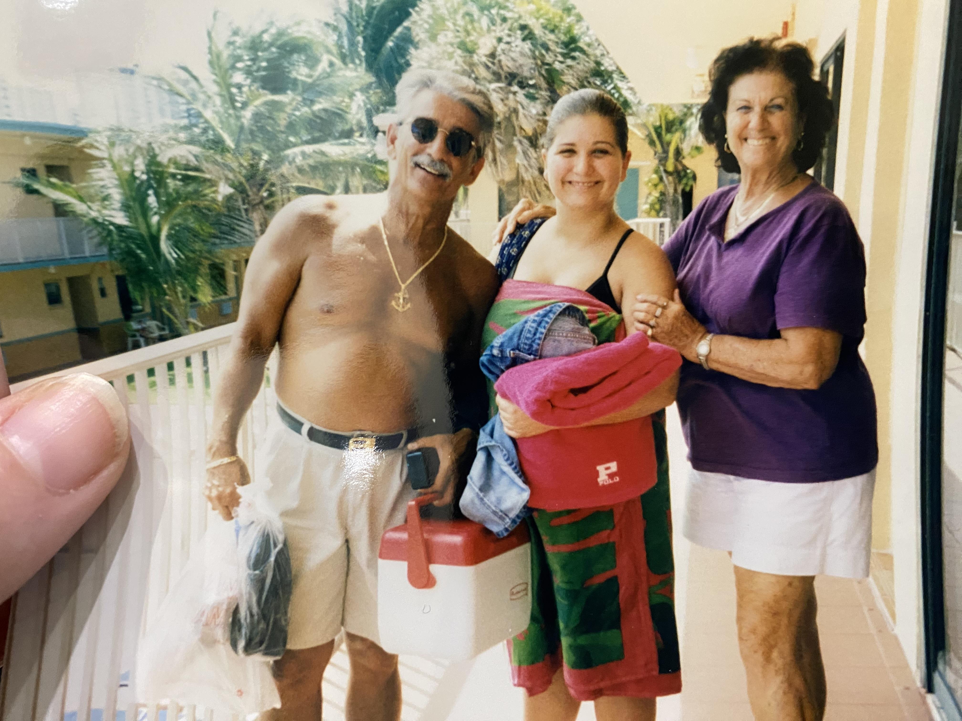 Three people smile on a balcony with beach gear, enjoying a sunny afternoon in paradise.