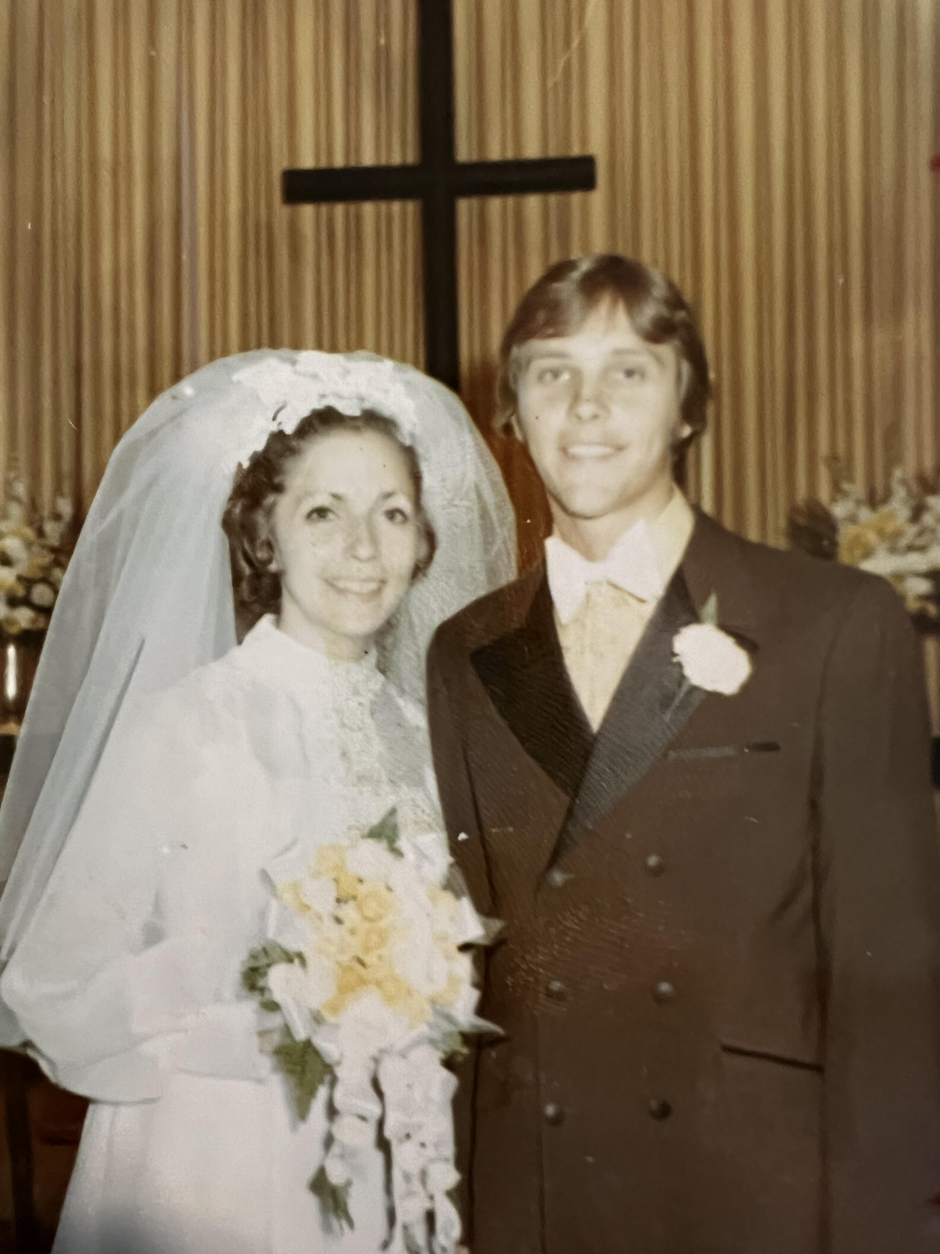 A happy couple in formal wedding attire smiles amid floral decorations.