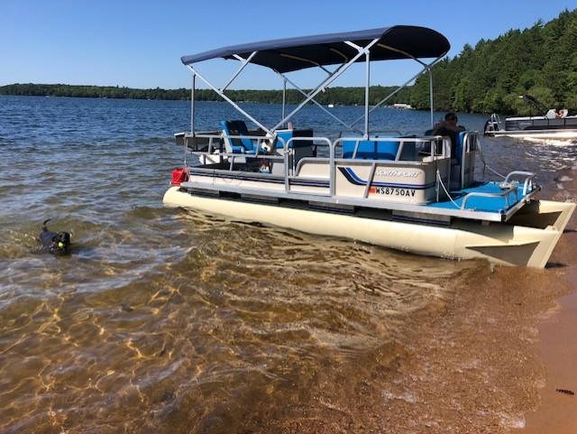 A pontoon boat is parked on the sandy beach as a dog plays in the water, enjoying the calm lake.