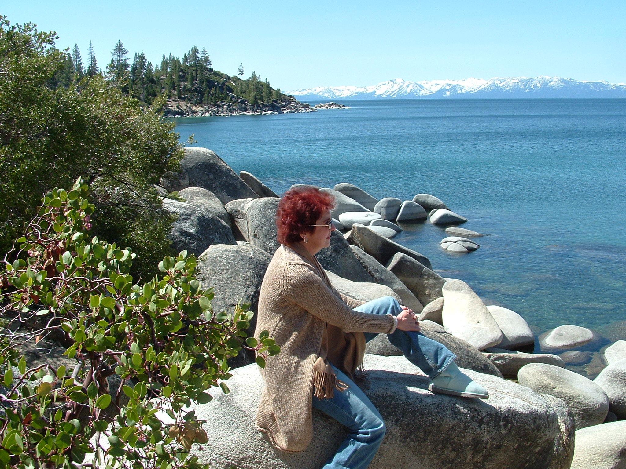 A woman enjoys the serene view of Lake Tahoe while sitting on large rocks under a clear sky.