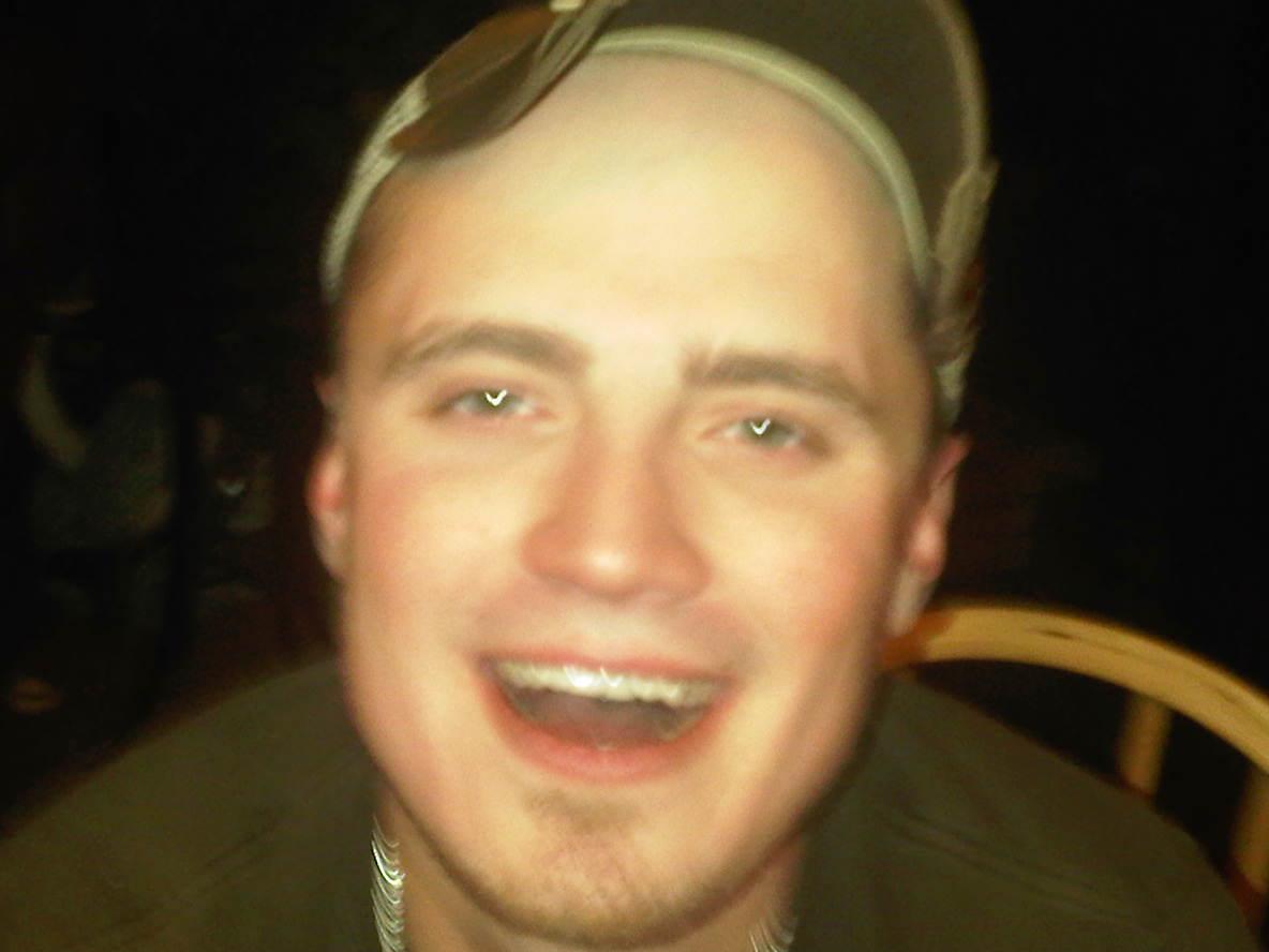 A young man with a joyful expression beams while wearing casual attire inside a cozy setting.