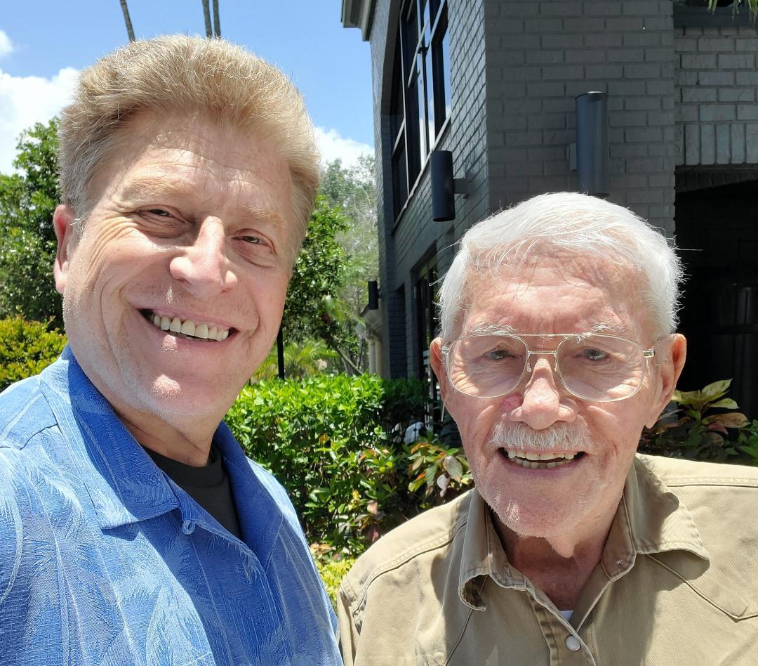 Two men pose happily for a selfie outside on a sunny day, surrounded by lush plants.