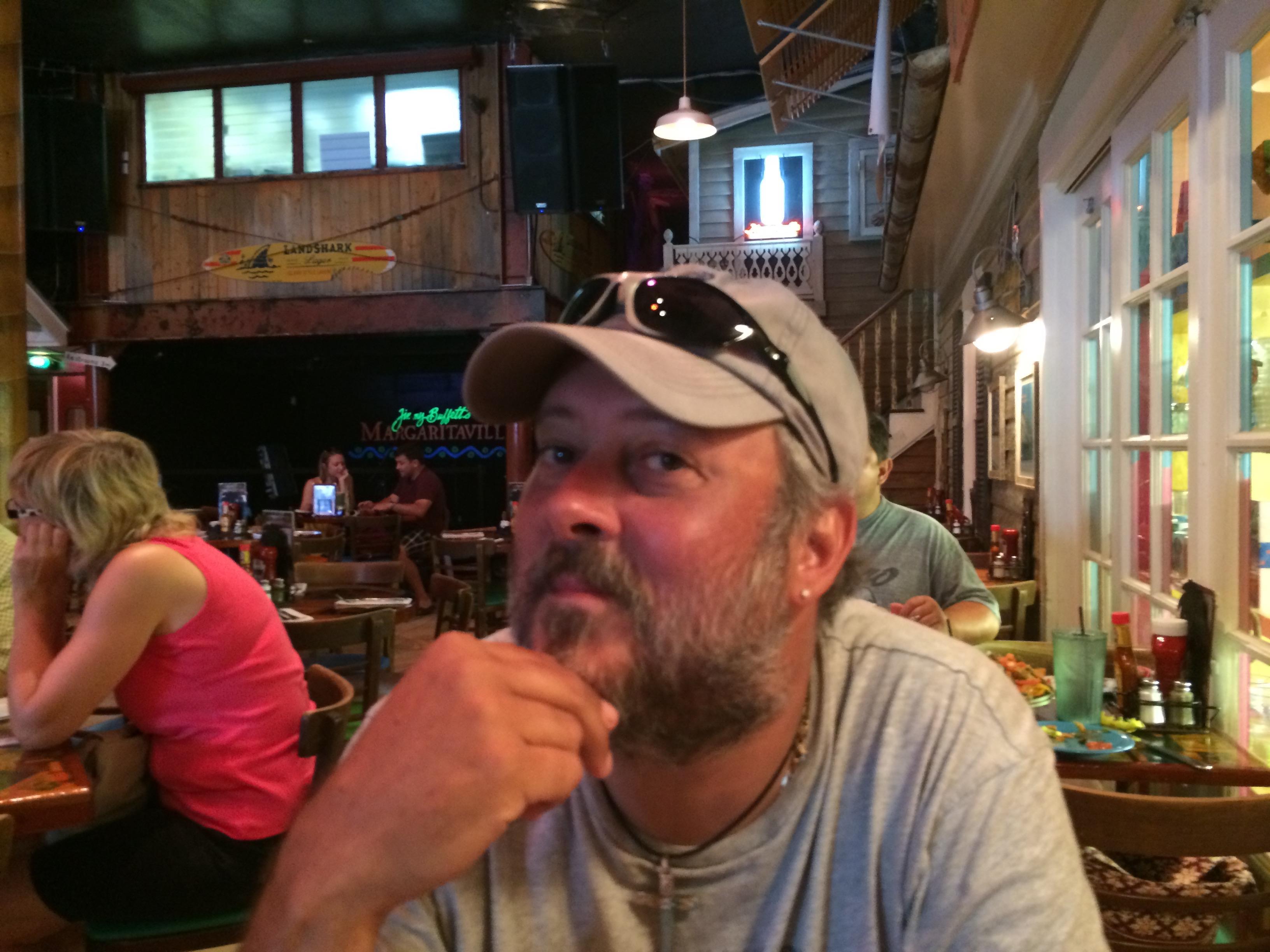A man sits at a table in a lively restaurant, smiling and deep in thought while others eat.