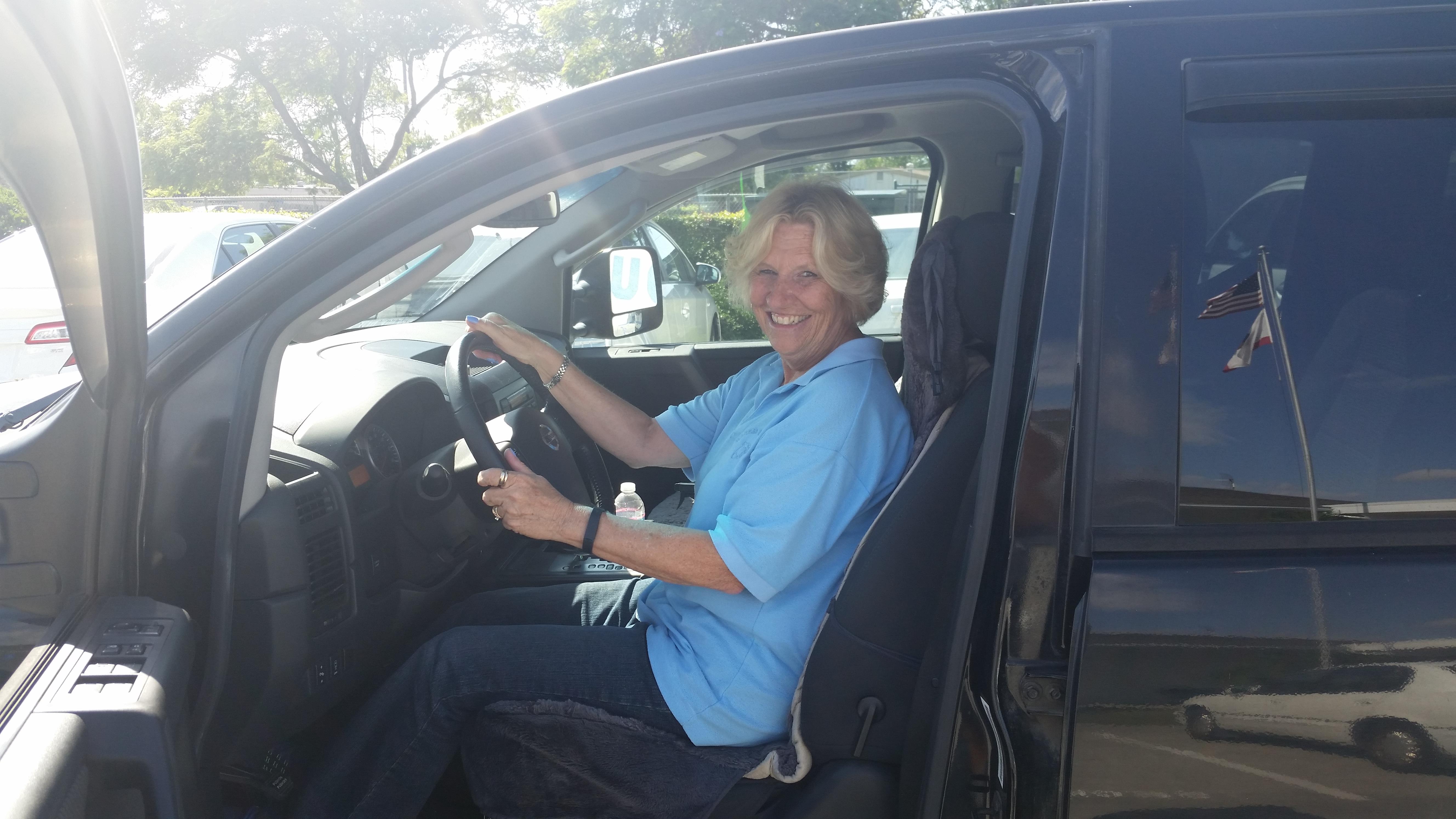 A smiling woman sits in the driver's seat of a black car parked in the sun.