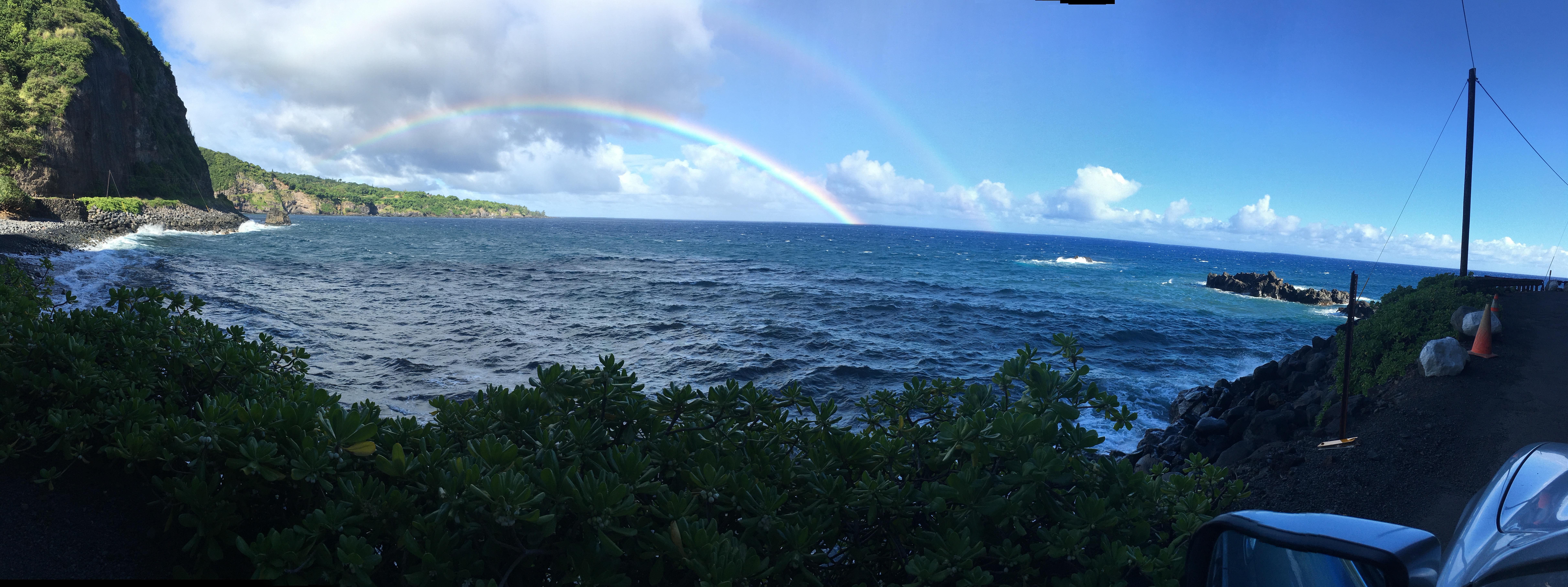 Ocean waves crash against rocky shorelines under a vibrant rainbow in a tropical landscape.