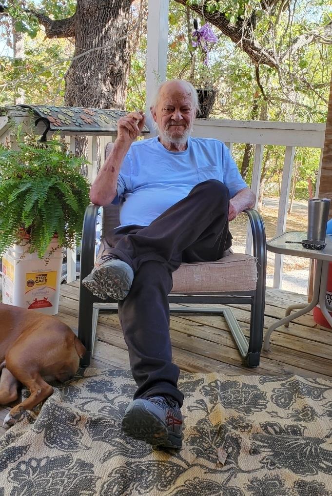 An older man sits comfortably on a porch, smoking while his dog rests nearby, surrounded by nature.
