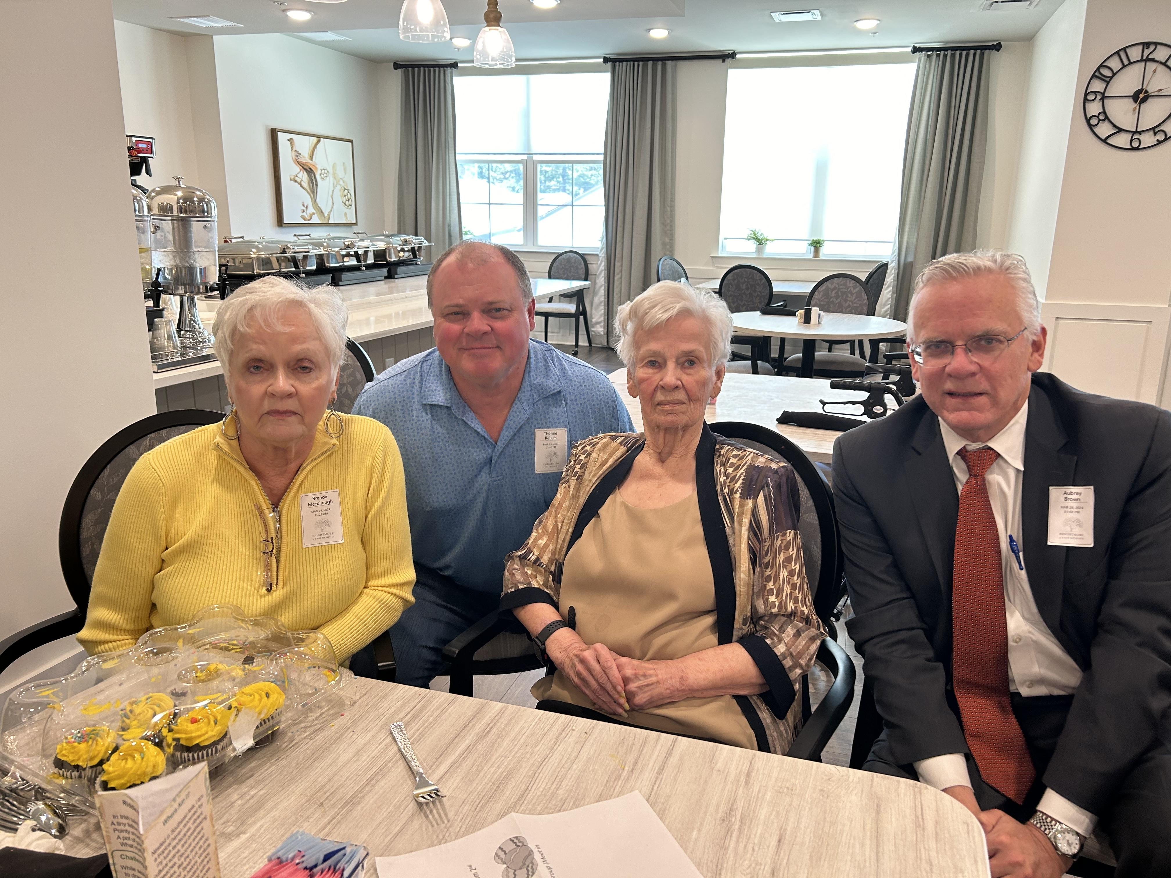 Four seniors enjoy a social gathering in a community hall with snacks and lively conversation.
