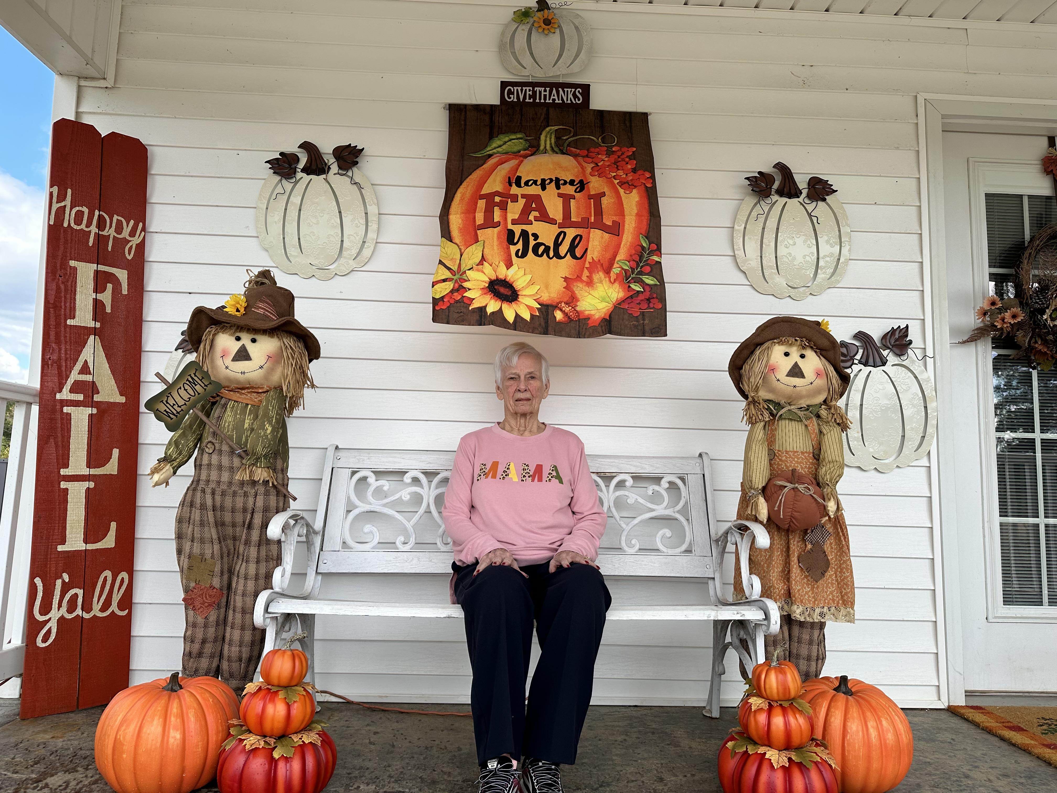 Elderly woman relaxes on porch surrounded by fall decor, pumpkins, and cheerful scarecrows.