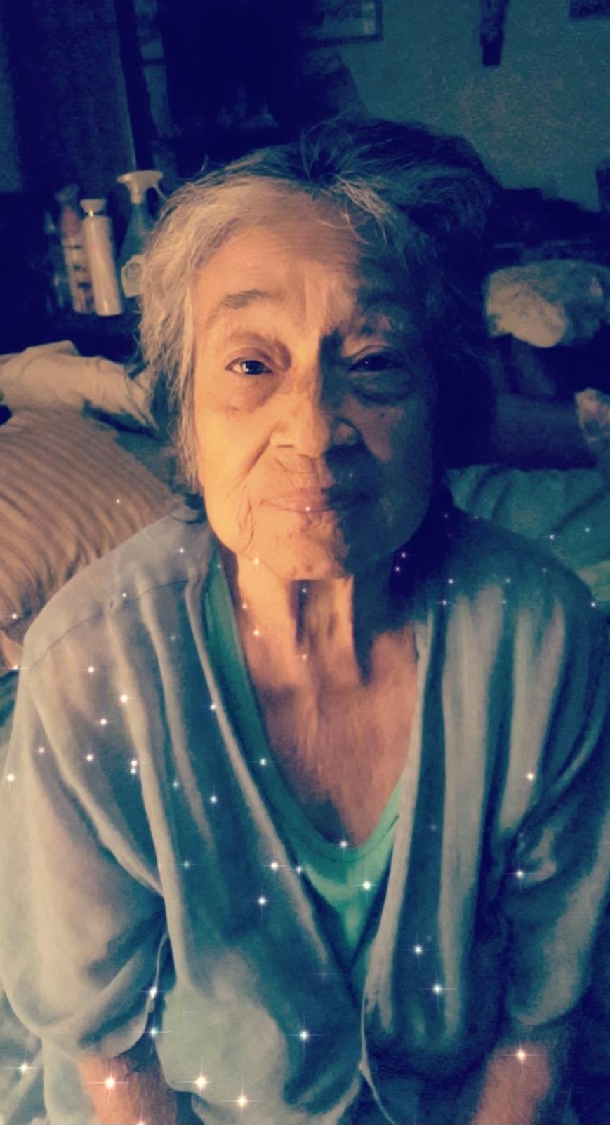 A joyful elderly woman is sitting on her bed, smiling gently at the camera in a dimly lit room.