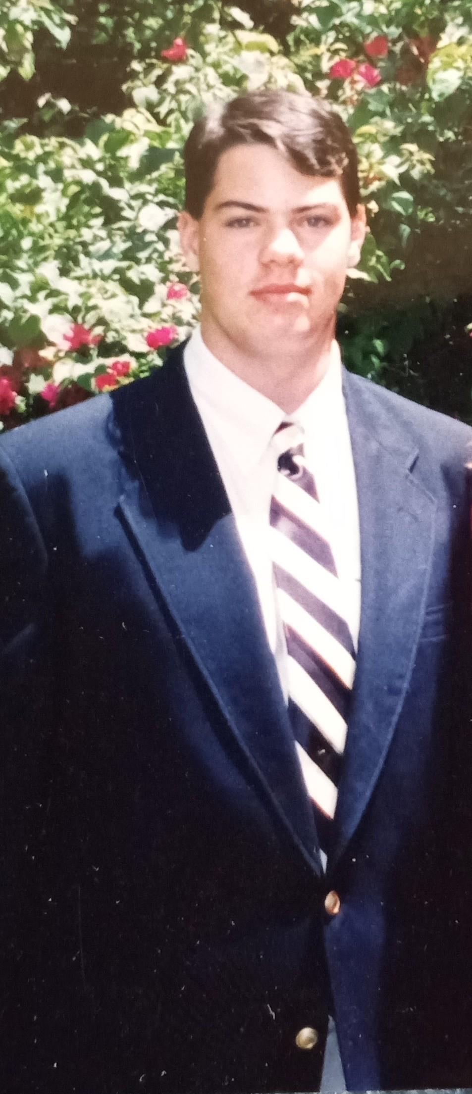 A man in a navy suit and striped tie stands confidently with flowers on a sunny day.