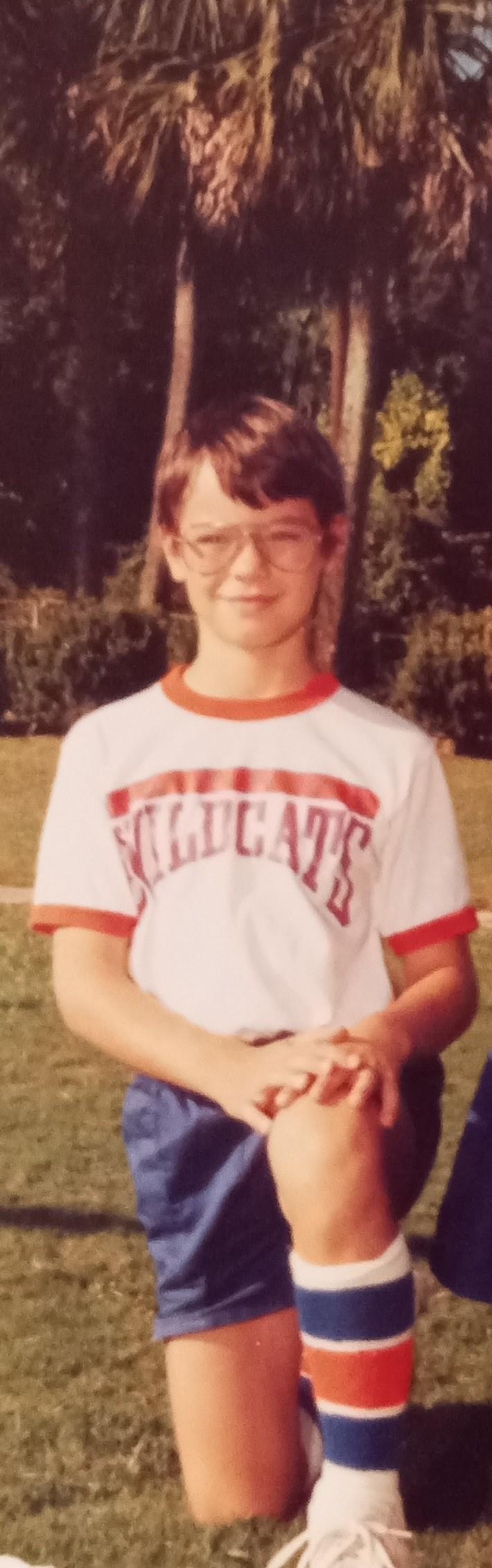A young boy stands confidently with a smile in a sports uniform on a grassy field.