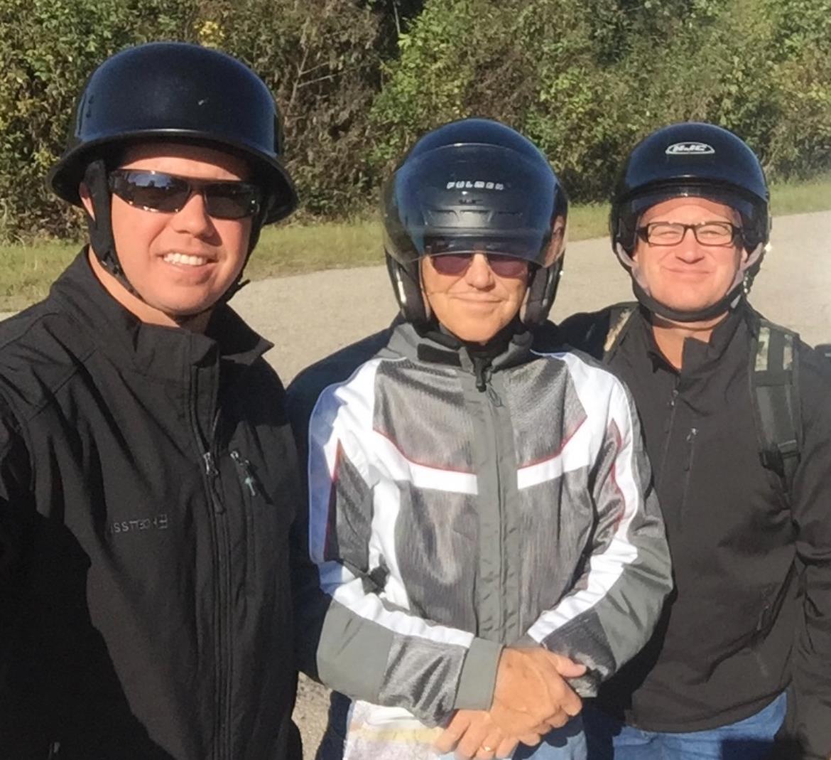 Three smiling motorcycle riders in helmets and jackets stand together in nature.