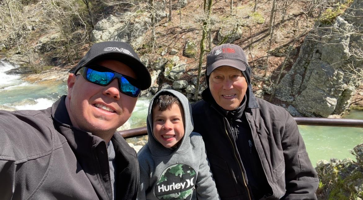 Three generations share smiles together along a beautiful river in a forested area.