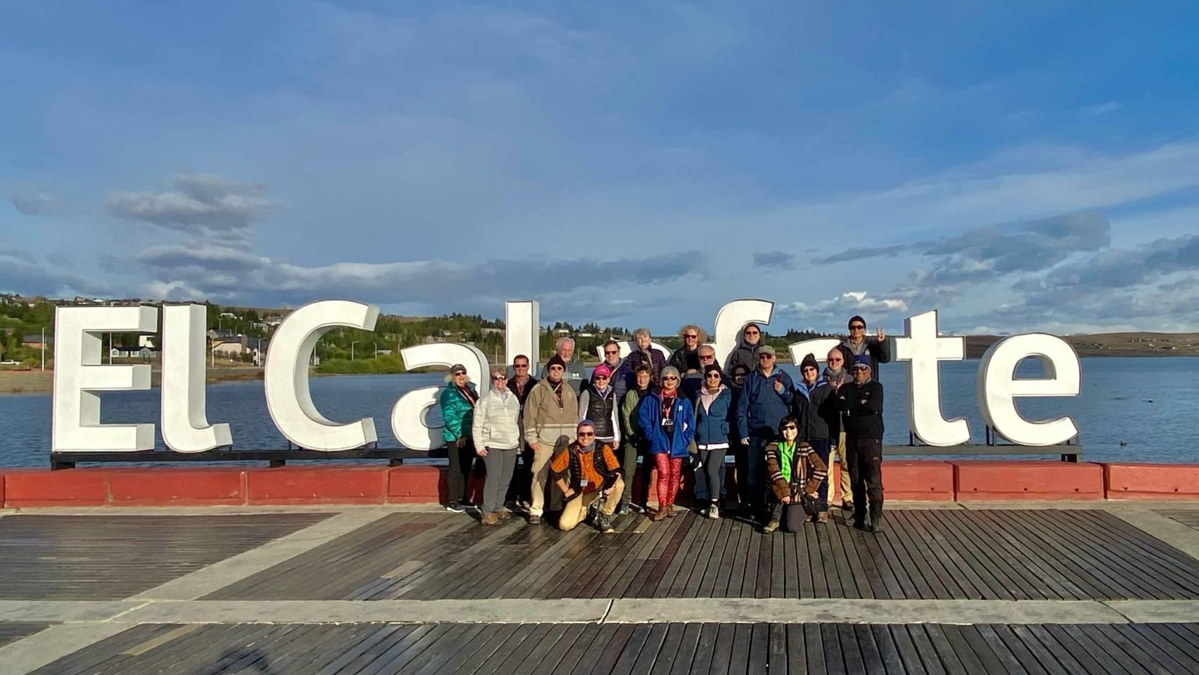 Tourists pose together in front of the El Calafate sign near a lake on a bright day in Patagonia.