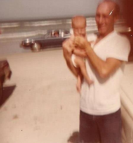 A man holds a baby near the shore on a warm day, with waves crashing and vintage cars parked nearby.