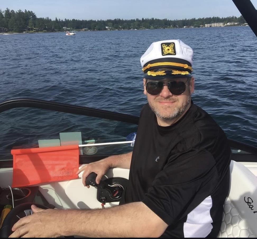 A man wears a captain's hat and sunglasses while steering a speedboat on a clear day.