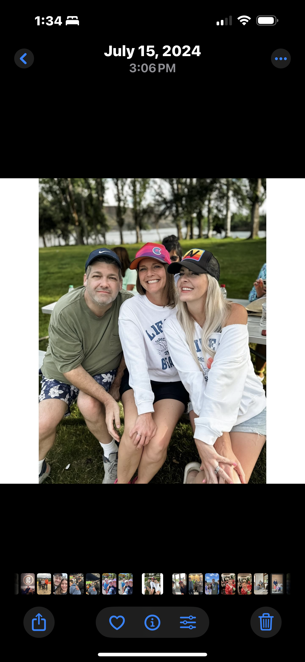 Three friends sit together in a park, wearing hats and laughing on a warm summer day by the water.