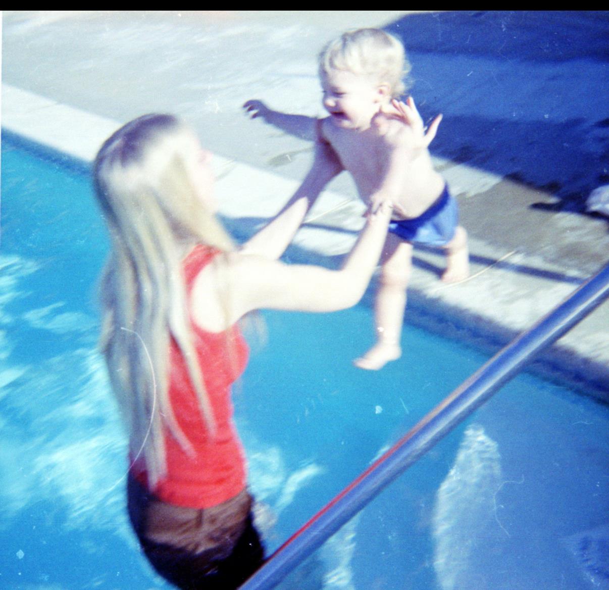 A woman plays with a child in a pool, lifting him up as he giggles with excitement on a sunny day.