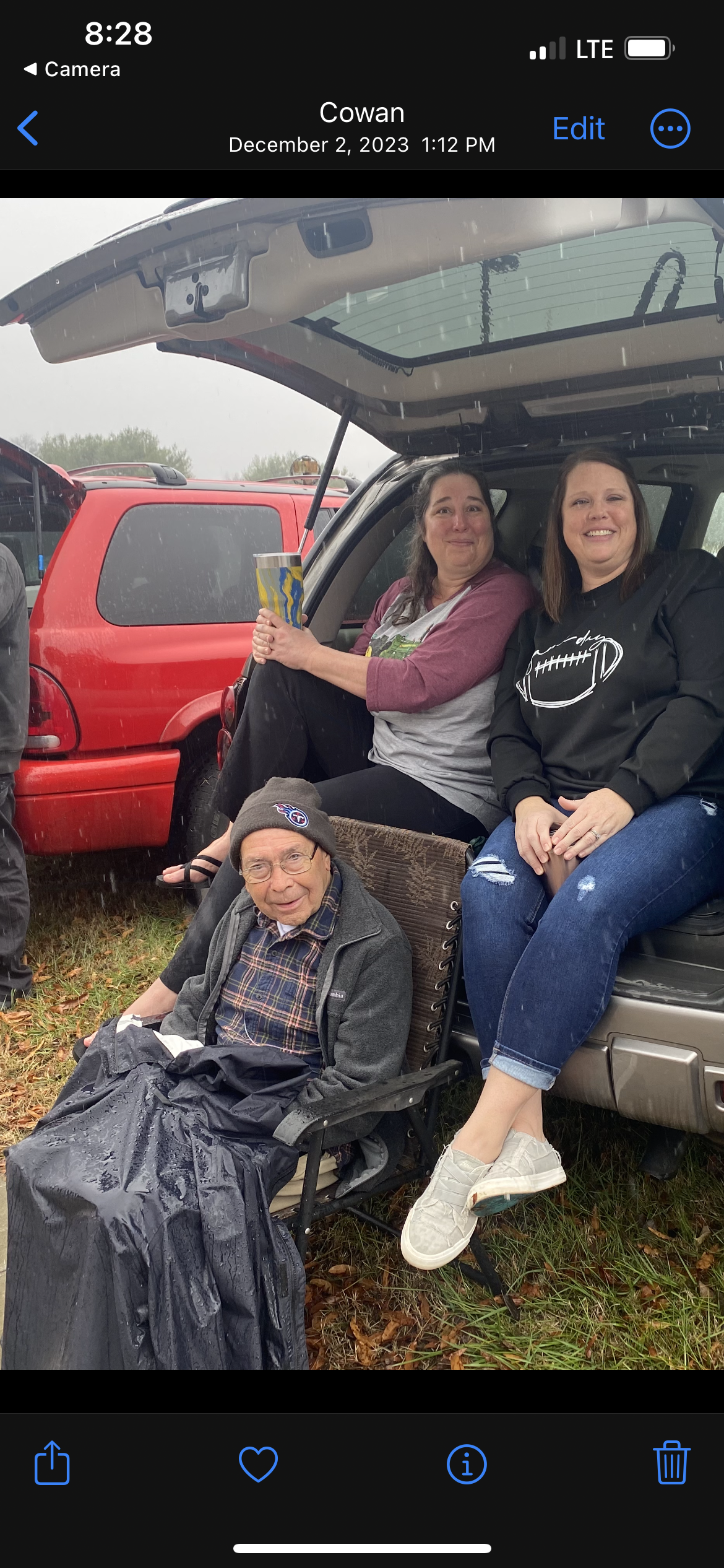 Three women relax in a parking lot while an elderly man sits in a chair, sheltered from rain.