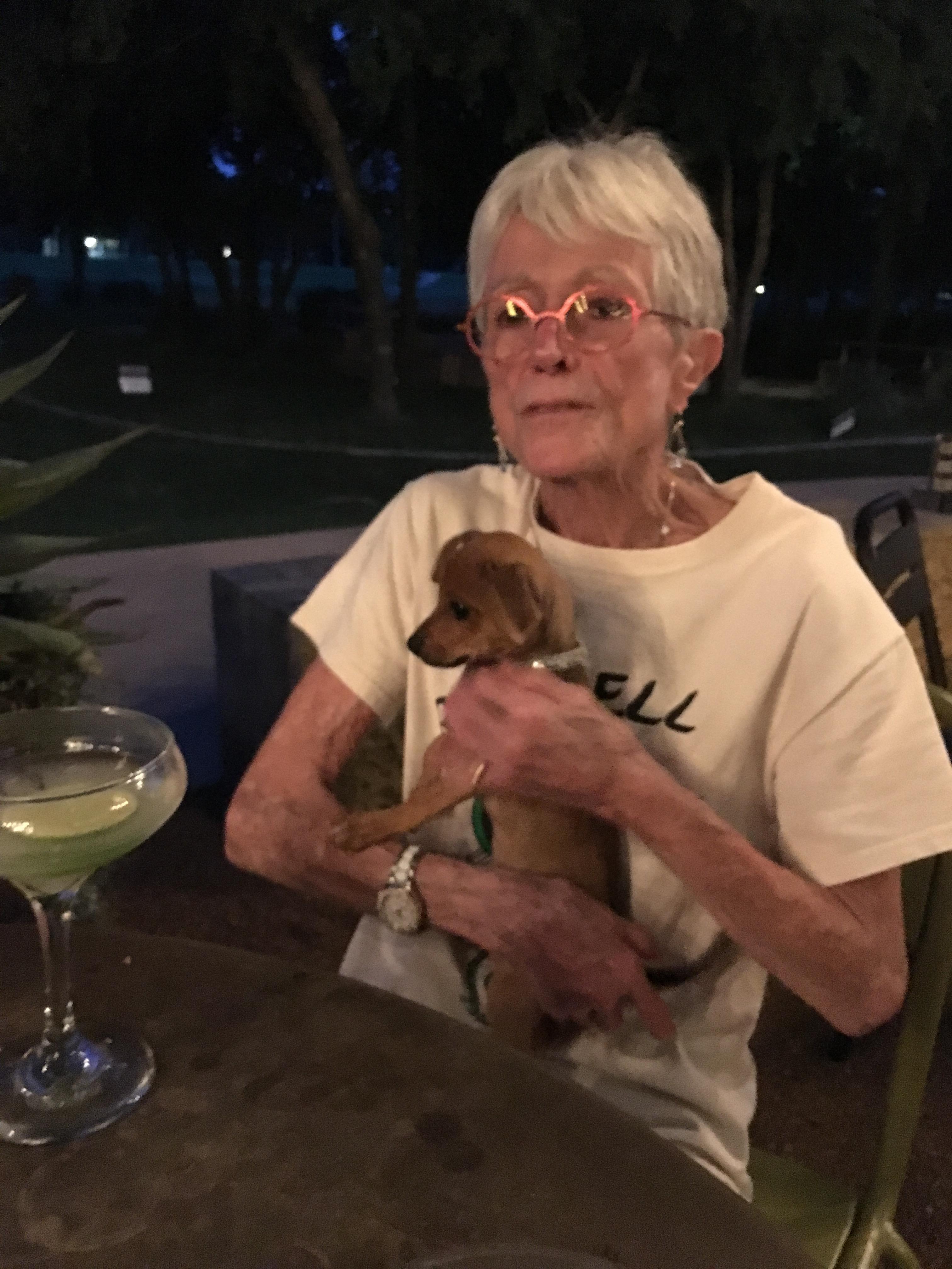 An older woman sits at a table, sipping a cocktail and cuddling a tiny puppy in the evening.