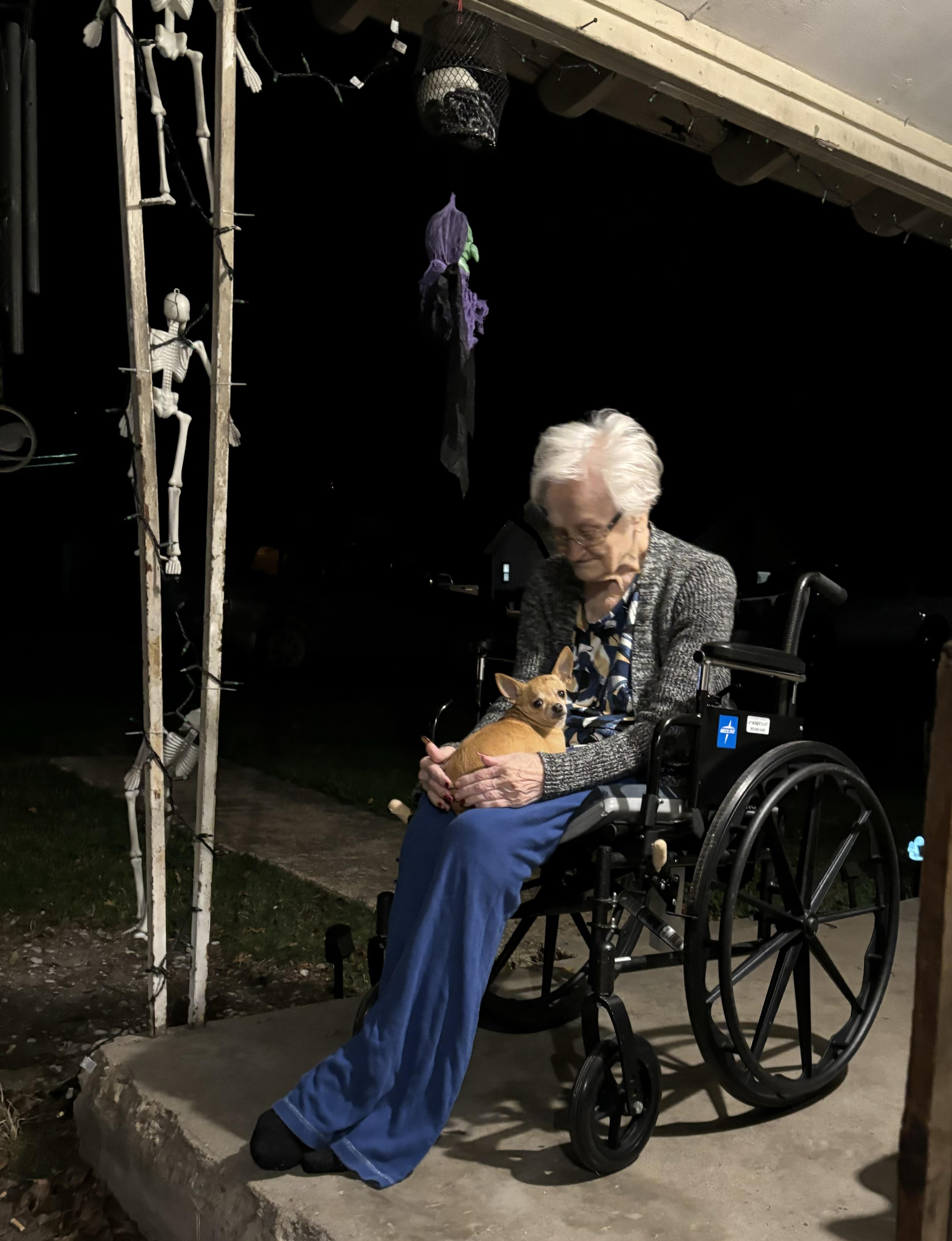 A woman in a wheelchair sits contently on a porch, holding a small dog while it’s dark outside.