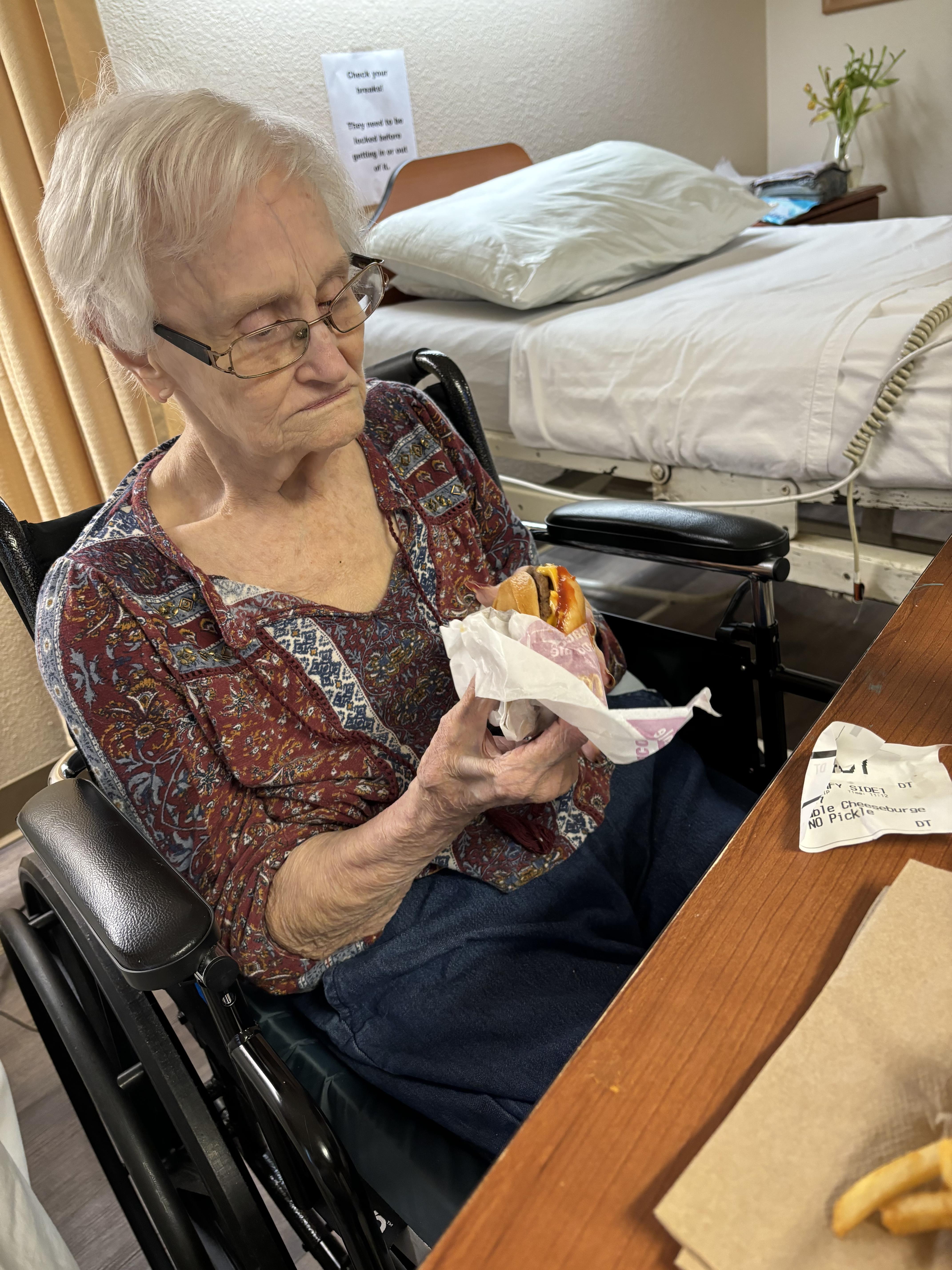Elderly woman in a wheelchair eats a sandwich at a care home, focused on her food.