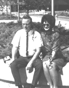 A man and woman sit together on a bench, wearing vintage attire, smiling under the sun.