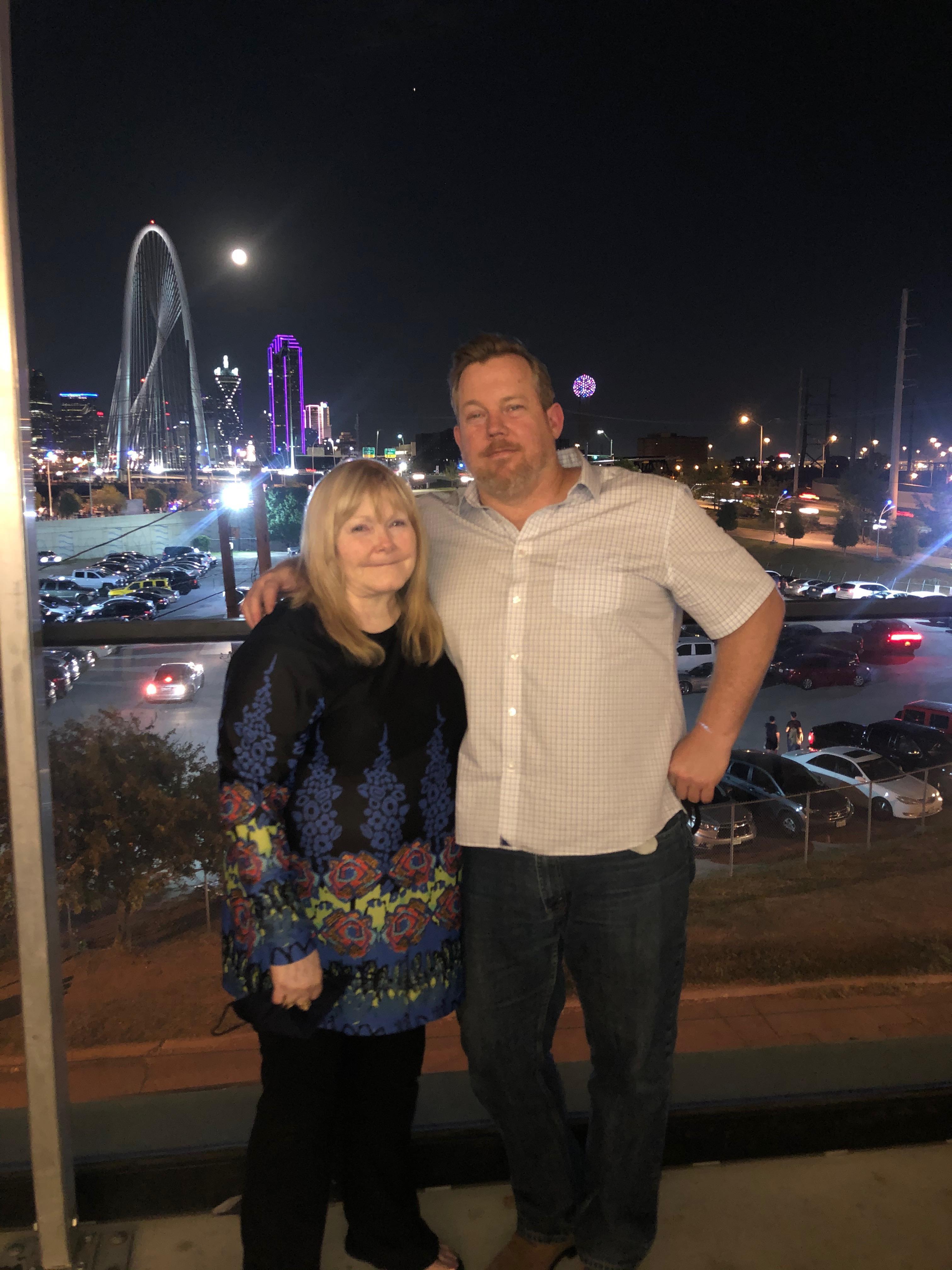 Two people pose joyfully at a city viewpoint with a glowing skyline behind them.