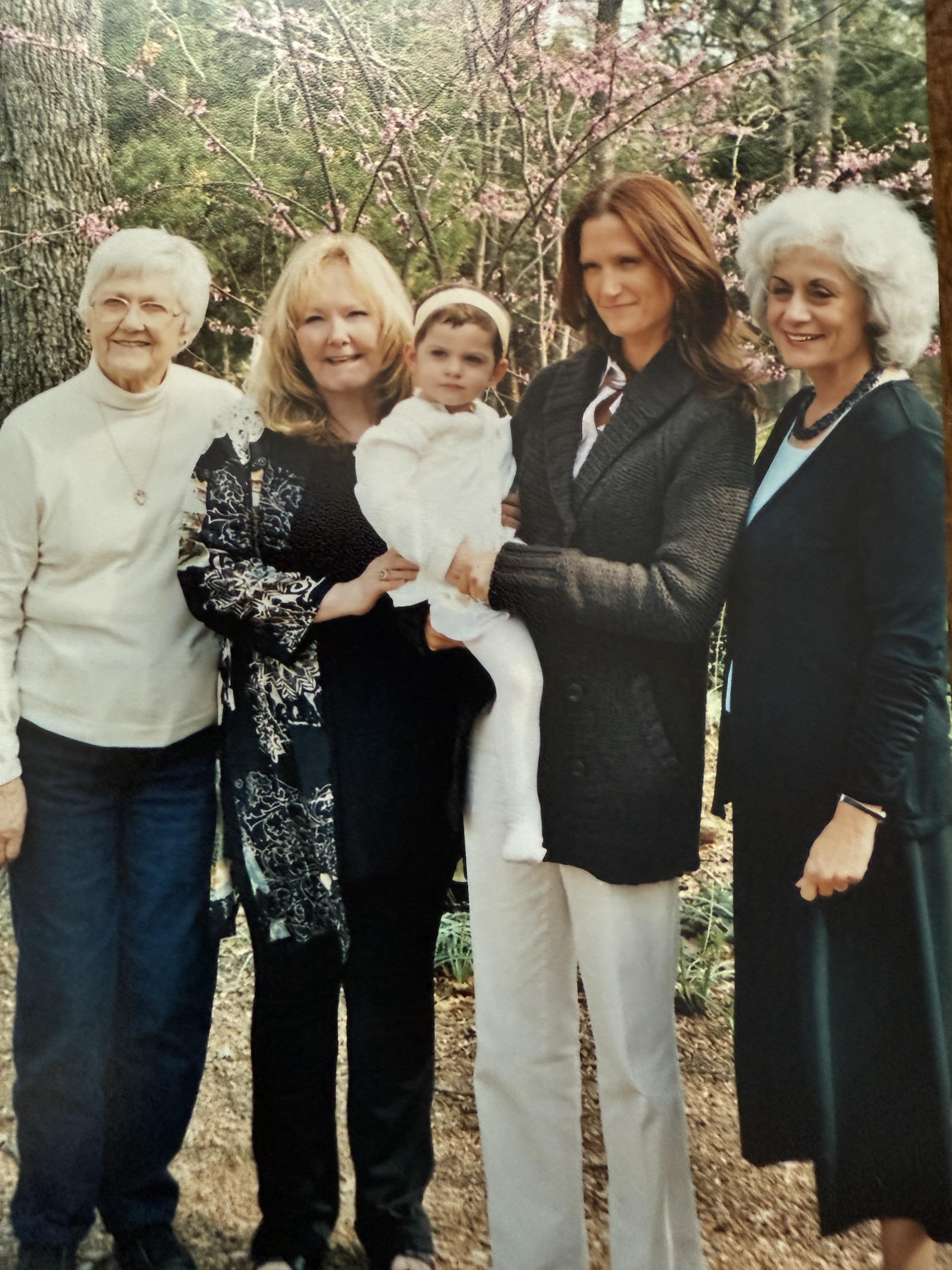 Four women of different generations gather in a garden holding a baby dressed in white.
