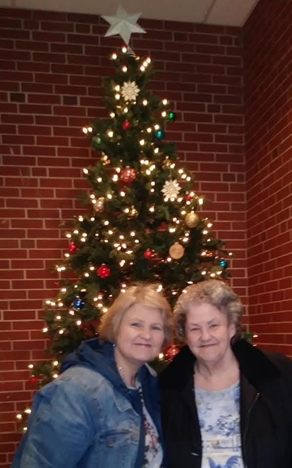 Two women smile happily while posing together by a beautifully decorated Christmas tree.