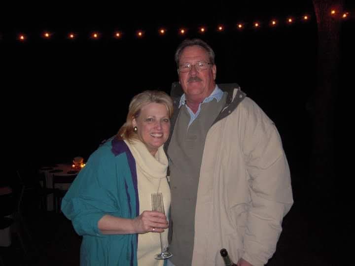 A couple smiles and poses together while holding drinks during a nighttime event.