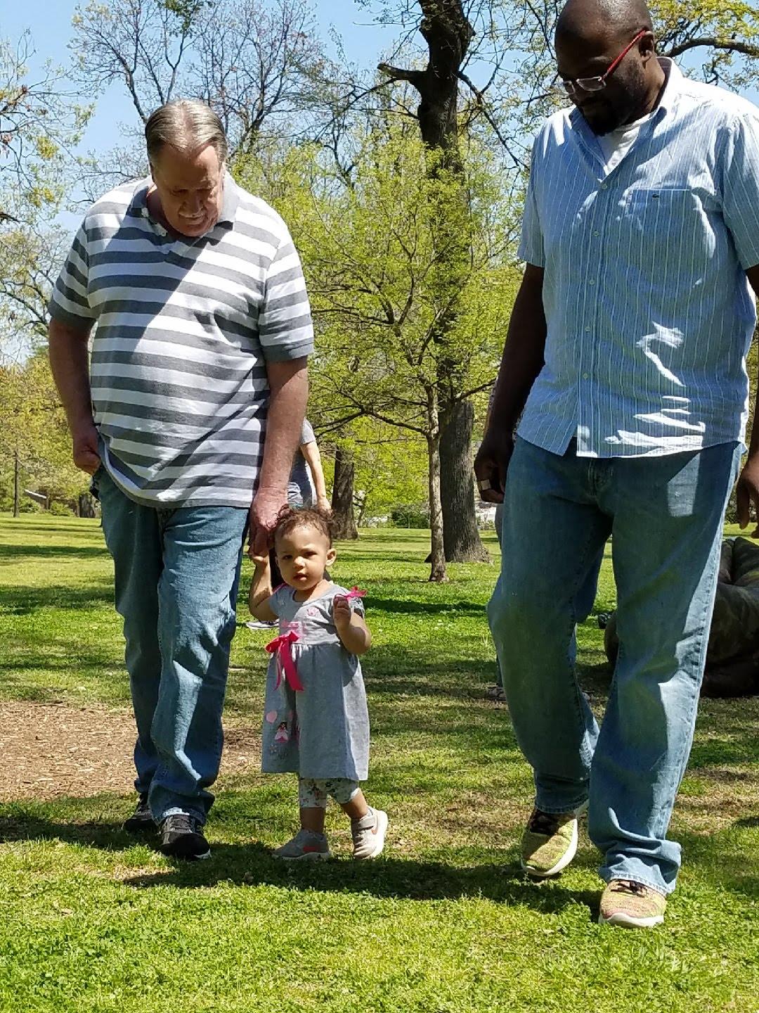 Two adults walk with a young child in a park filled with trees on a clear sunny day.