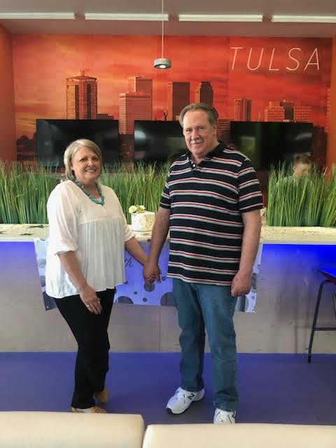 A couple is smiling and holding hands at a modern indoor space in Tulsa, surrounded by greenery.