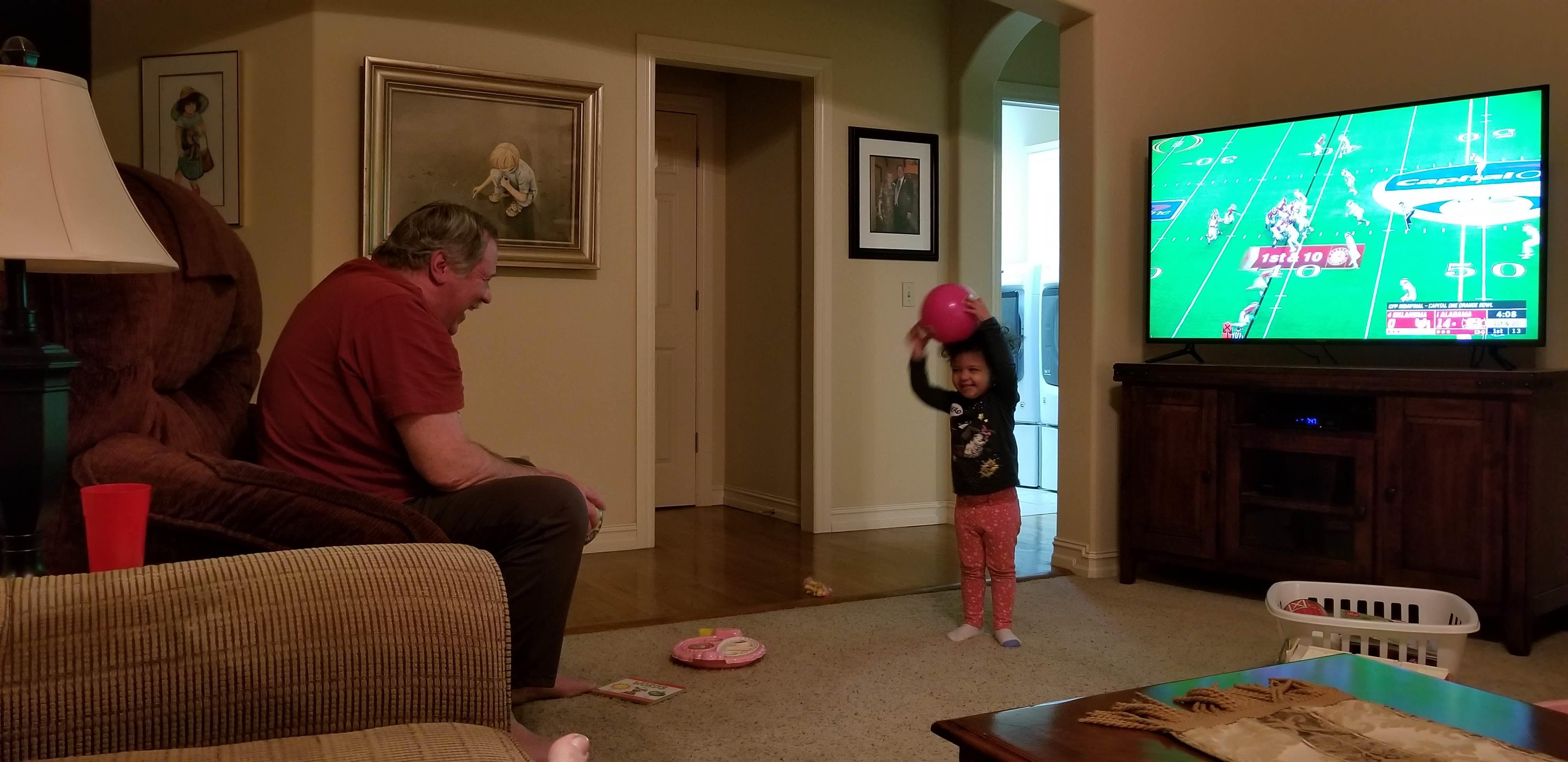 A young girl plays with a balloon while her grandfather watches a football game at home.