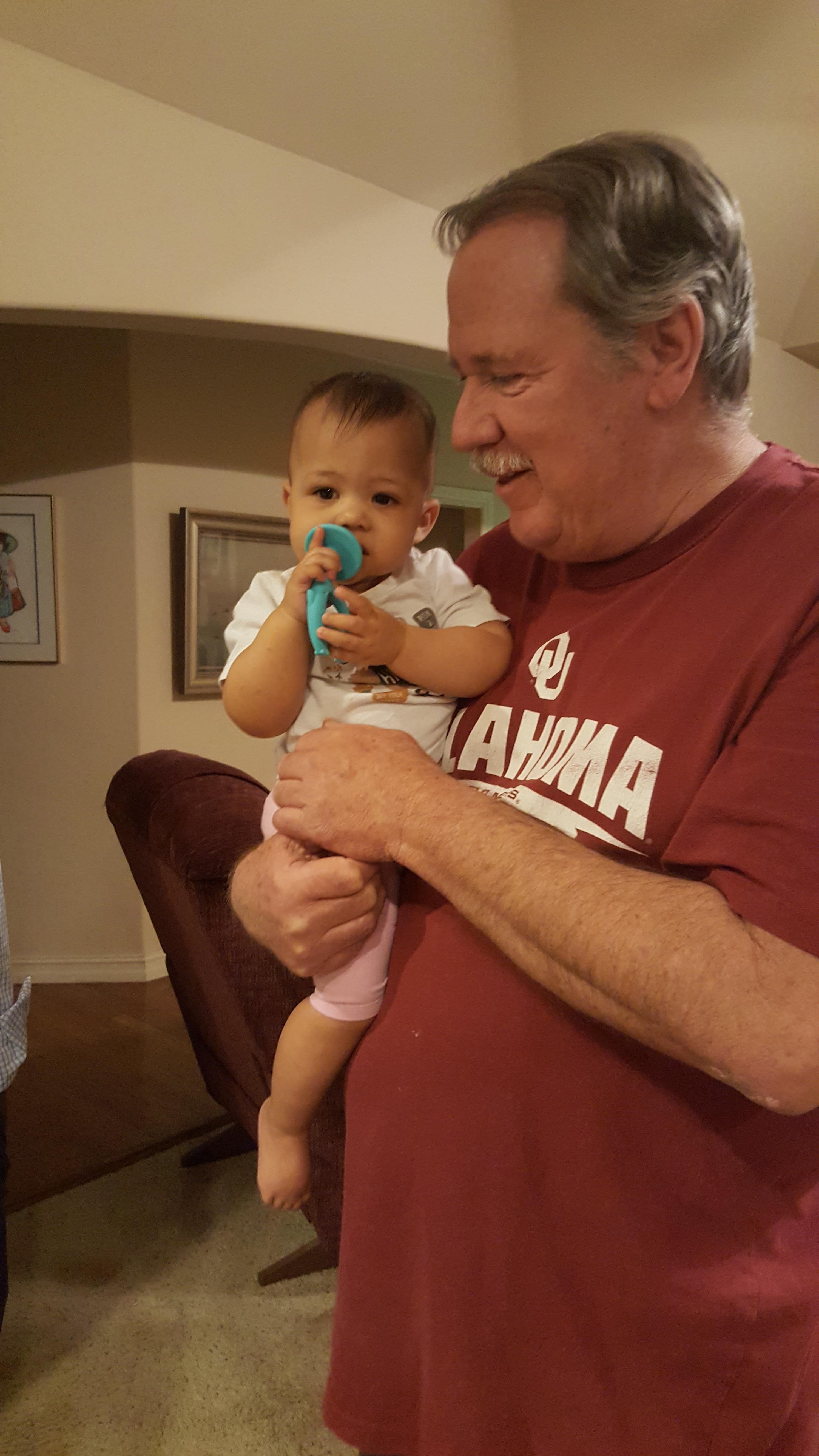 A joyful grandfather holds his baby granddaughter, who enjoys chewing on a toy at home.