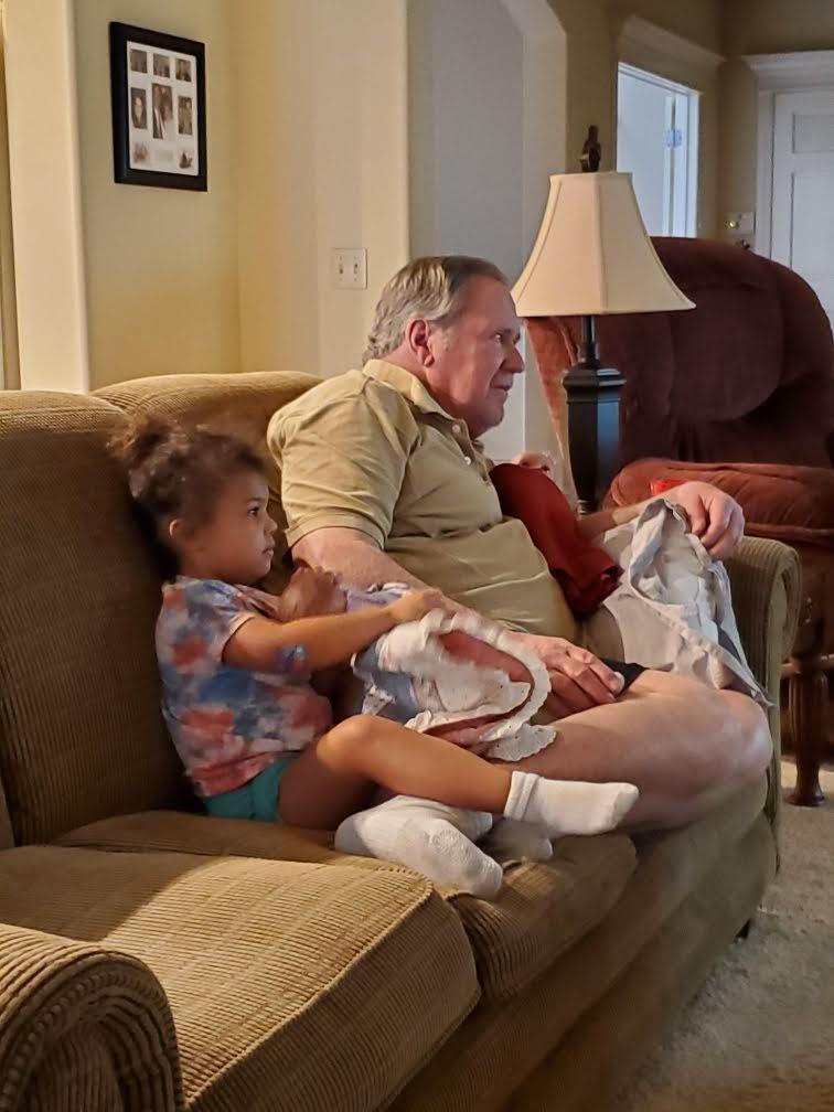 A young girl sits beside her grandfather, both relaxed, enjoying a moment on a comfortable couch.