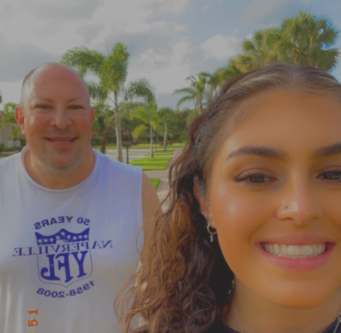 Two friends smile widely while taking a selfie, surrounded by lush greenery and sunshine.