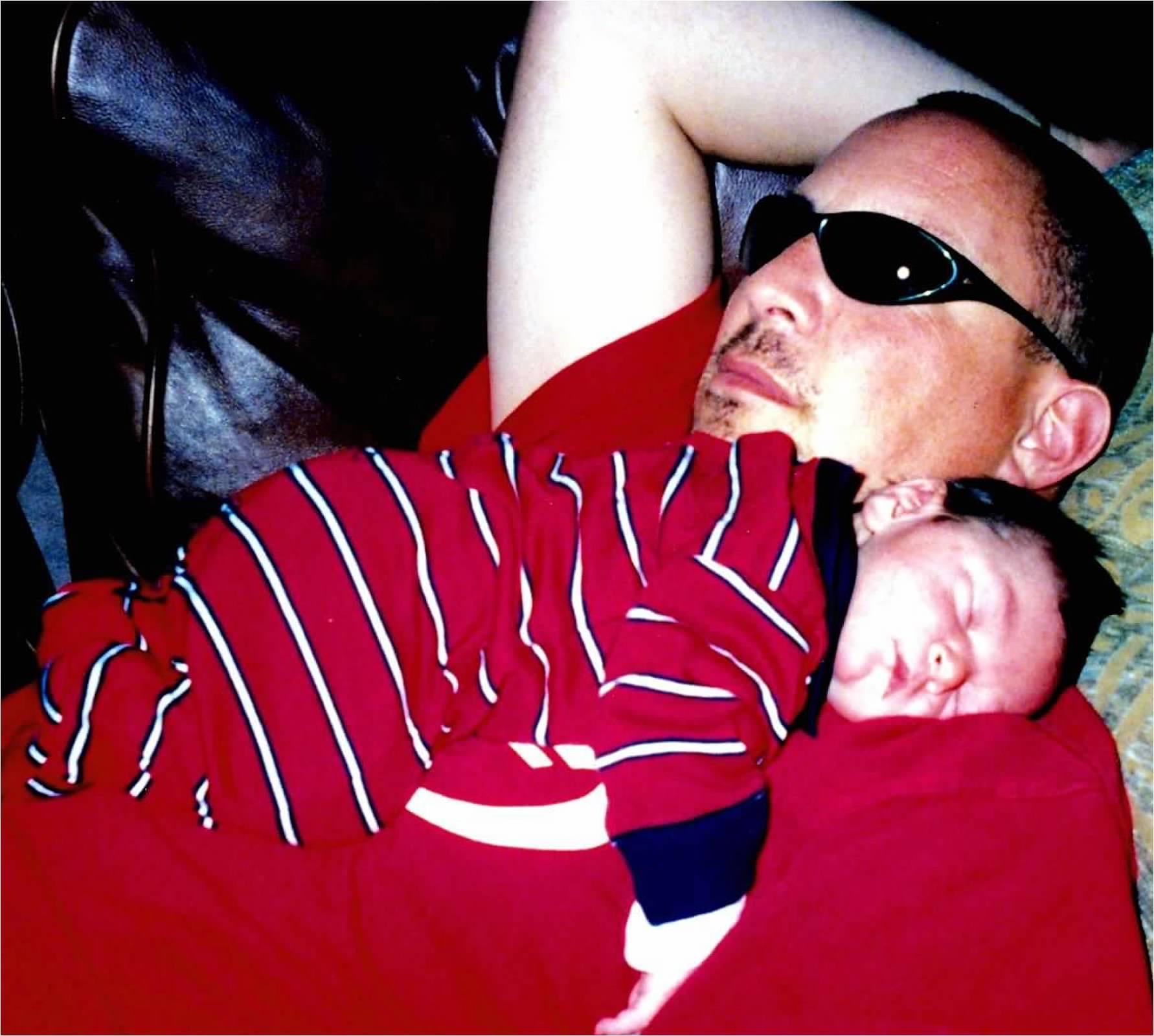 A father relaxes on a couch, sunglasses on, as his baby peacefully sleeps on his chest.
