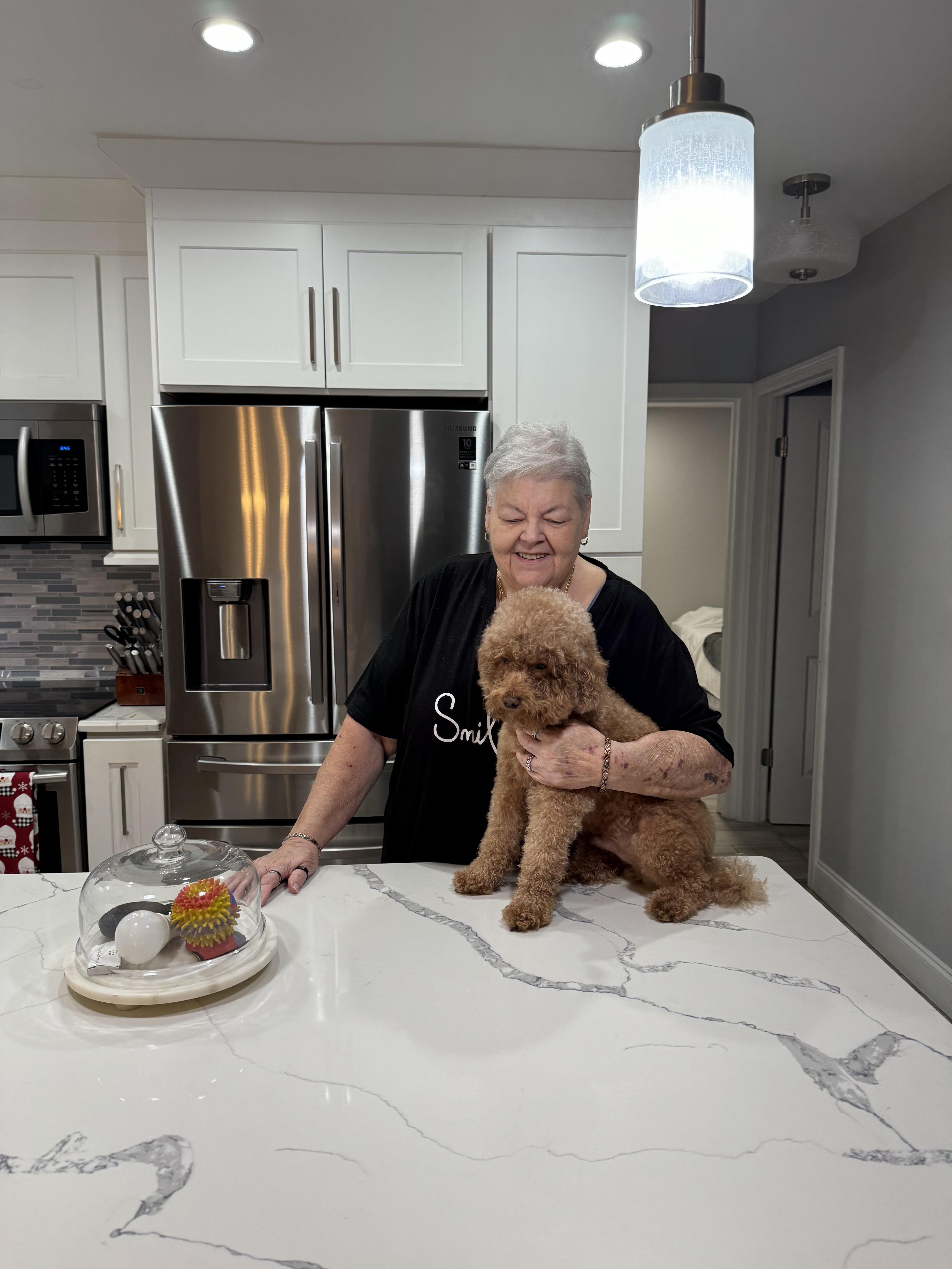 An elderly woman smiles warmly while holding her dog in a stylish kitchen setting, enjoying the day.