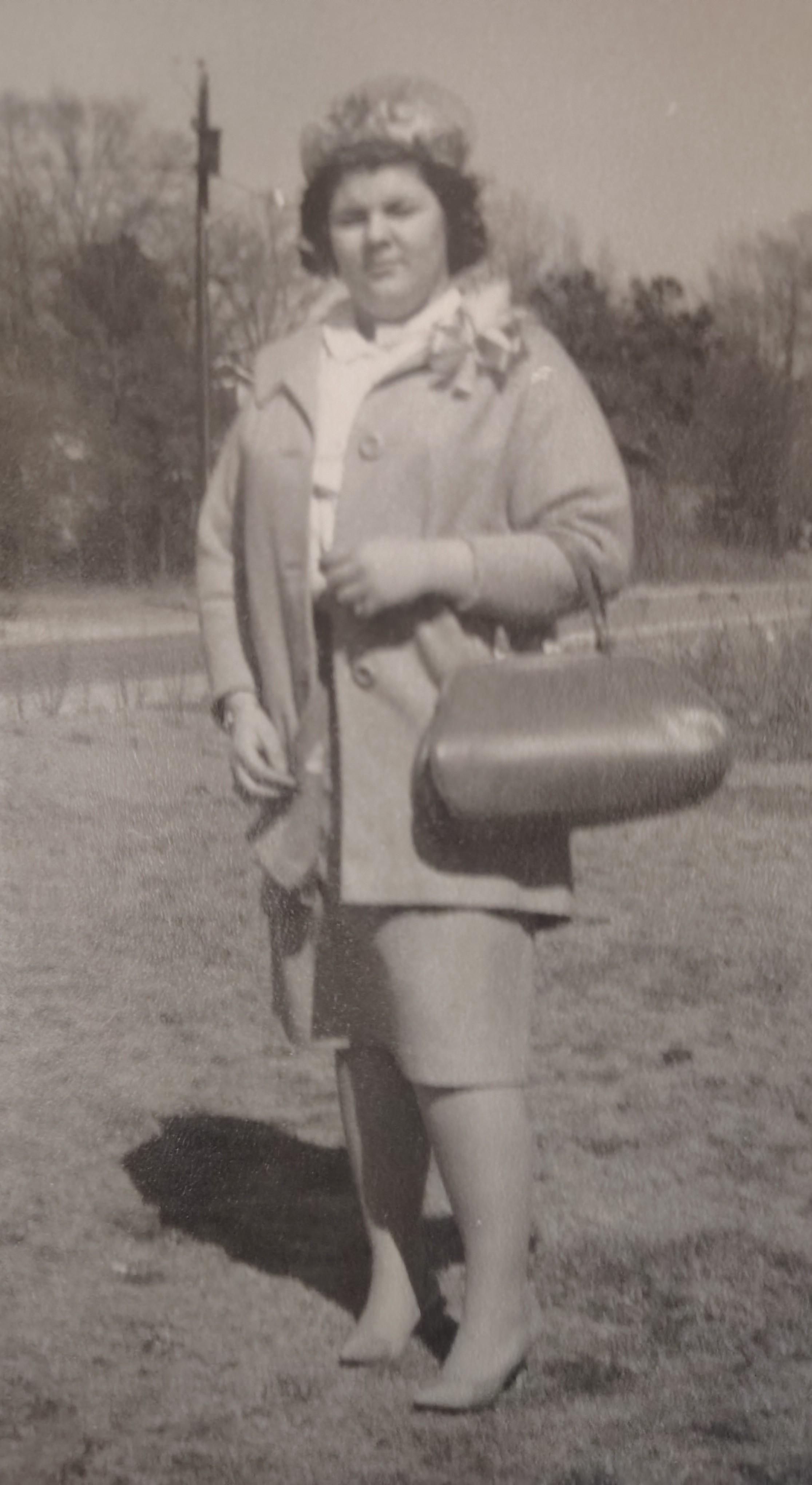 A woman in a stylish coat and heels confidently holds a handbag in rural spring.