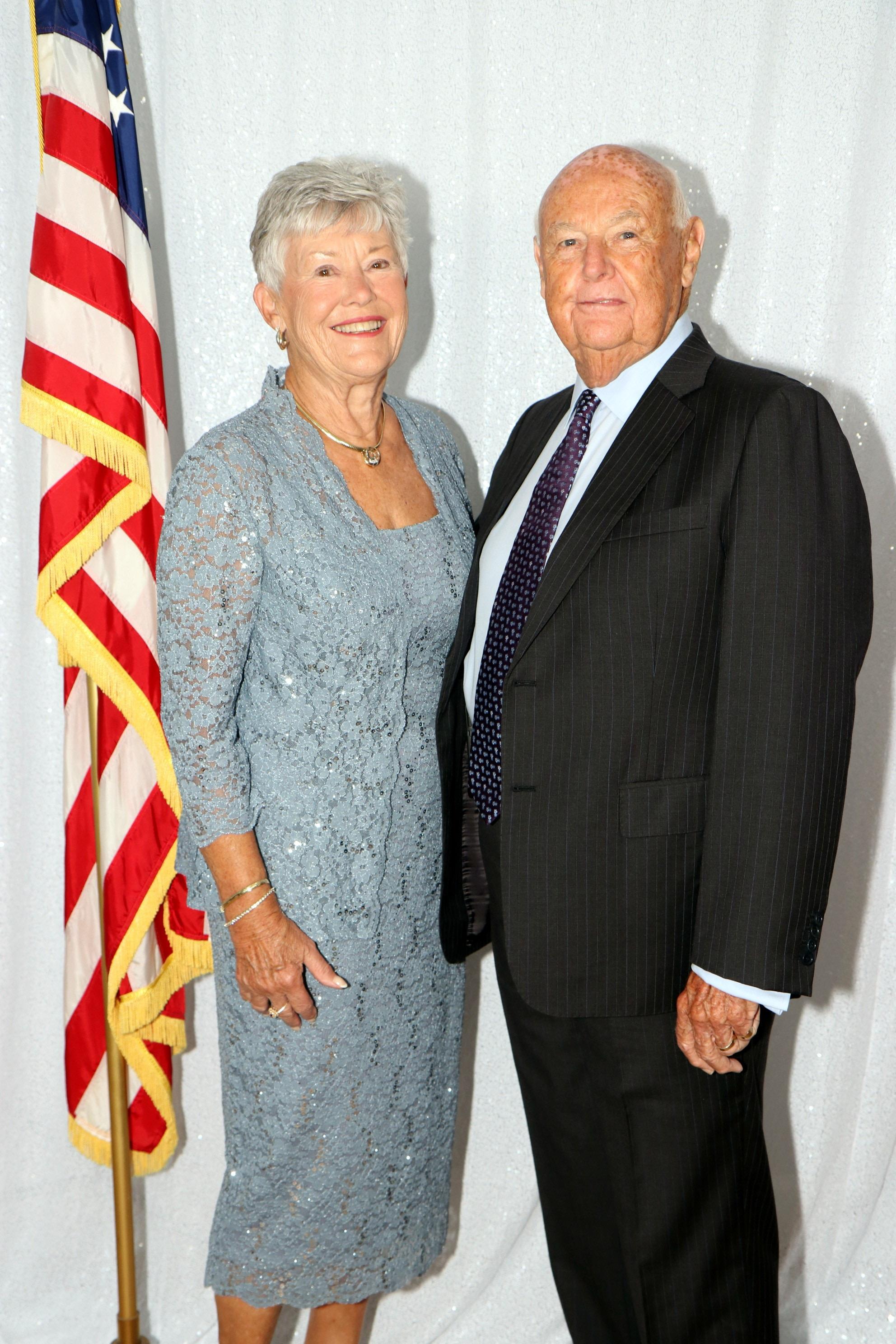 Dressed in formal attire, a couple smiles together against a backdrop featuring an American flag.