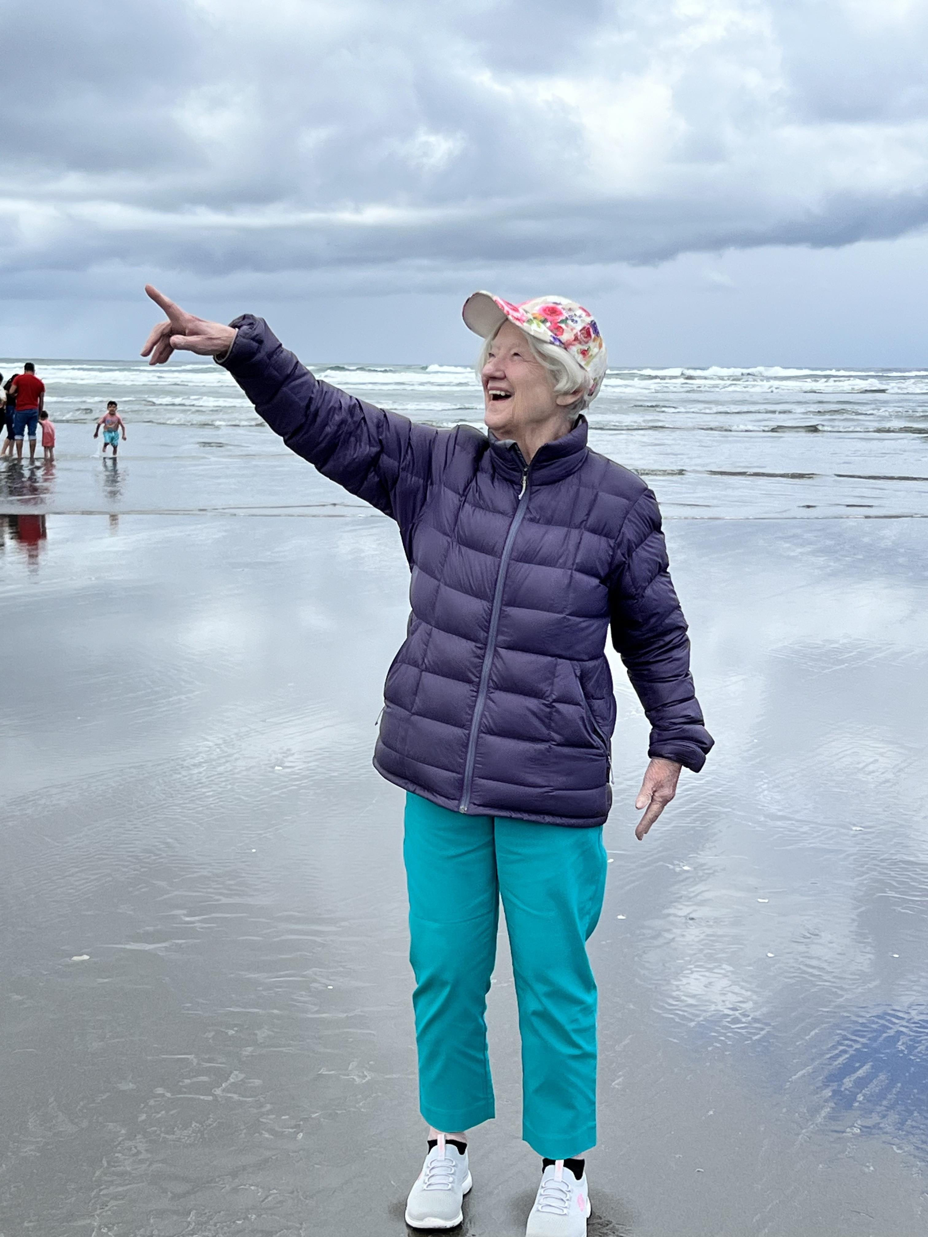 An elderly woman in a jacket and hat joyfully points at the ocean, enjoying a breezy beach day.