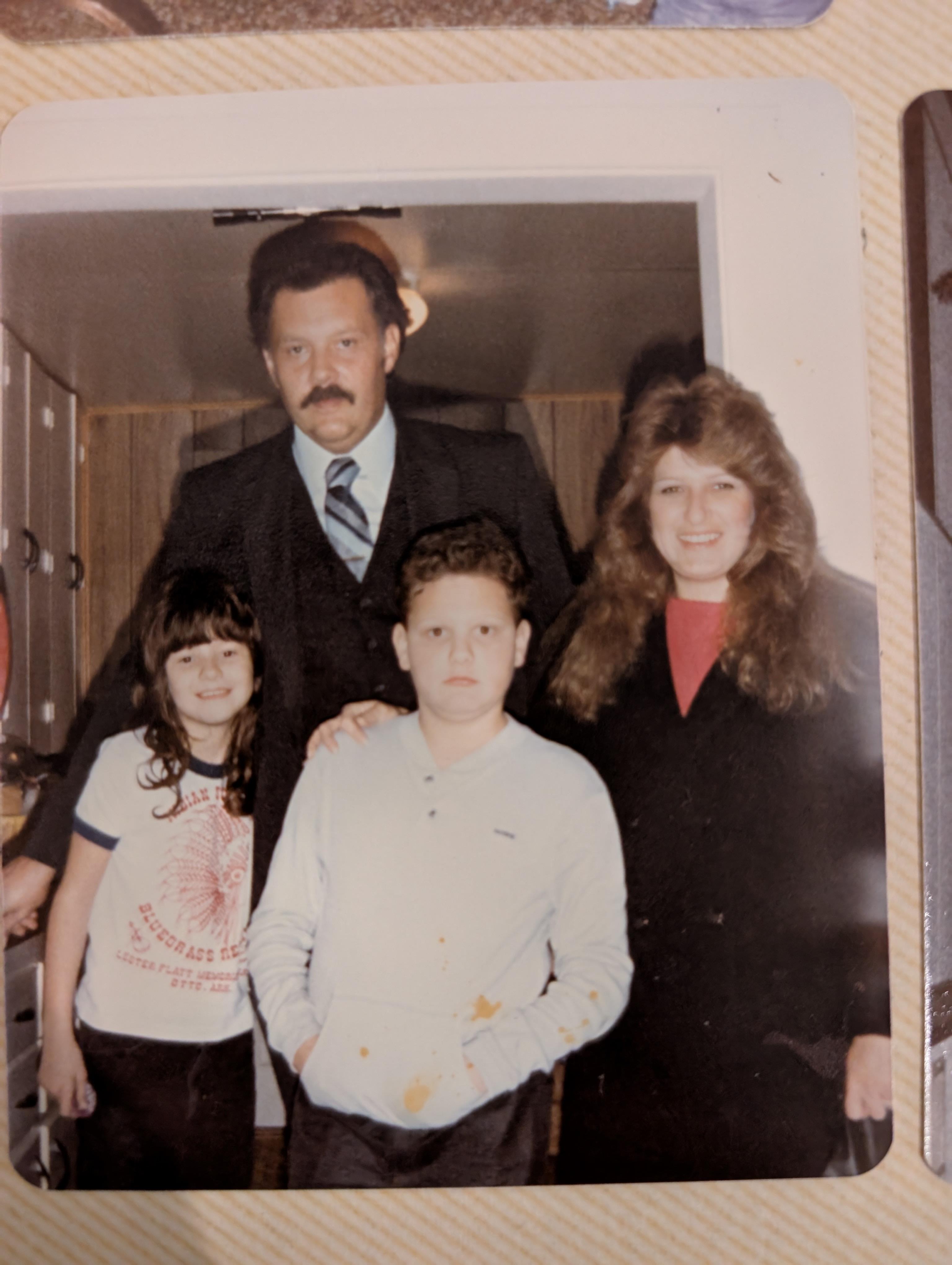 A family stands together in a cozy indoor setting, dressed in formal attire, captured in the 1980s.