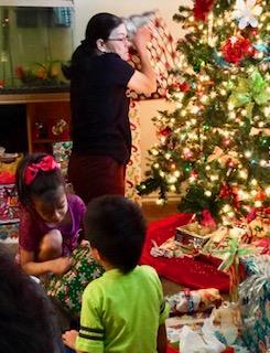 Children excitedly open presents while adults prepare decorations in a cozy living room setting.