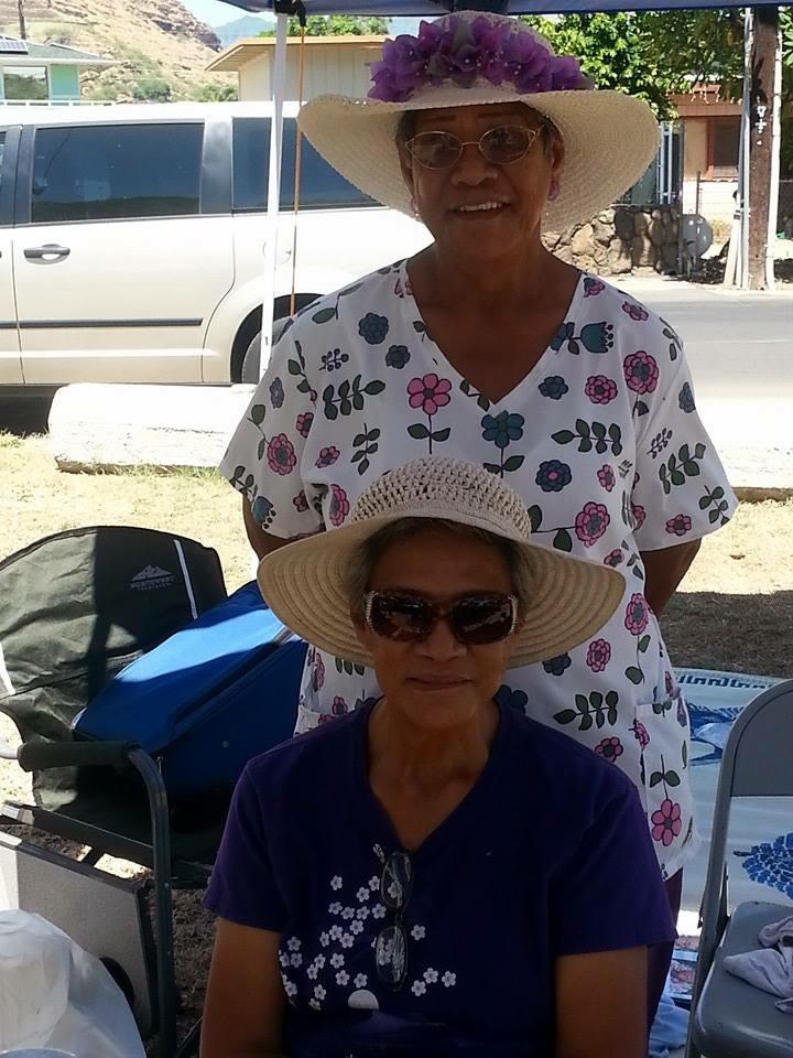 Two women are sitting together in hats at an outdoor market. They are selling handmade items.