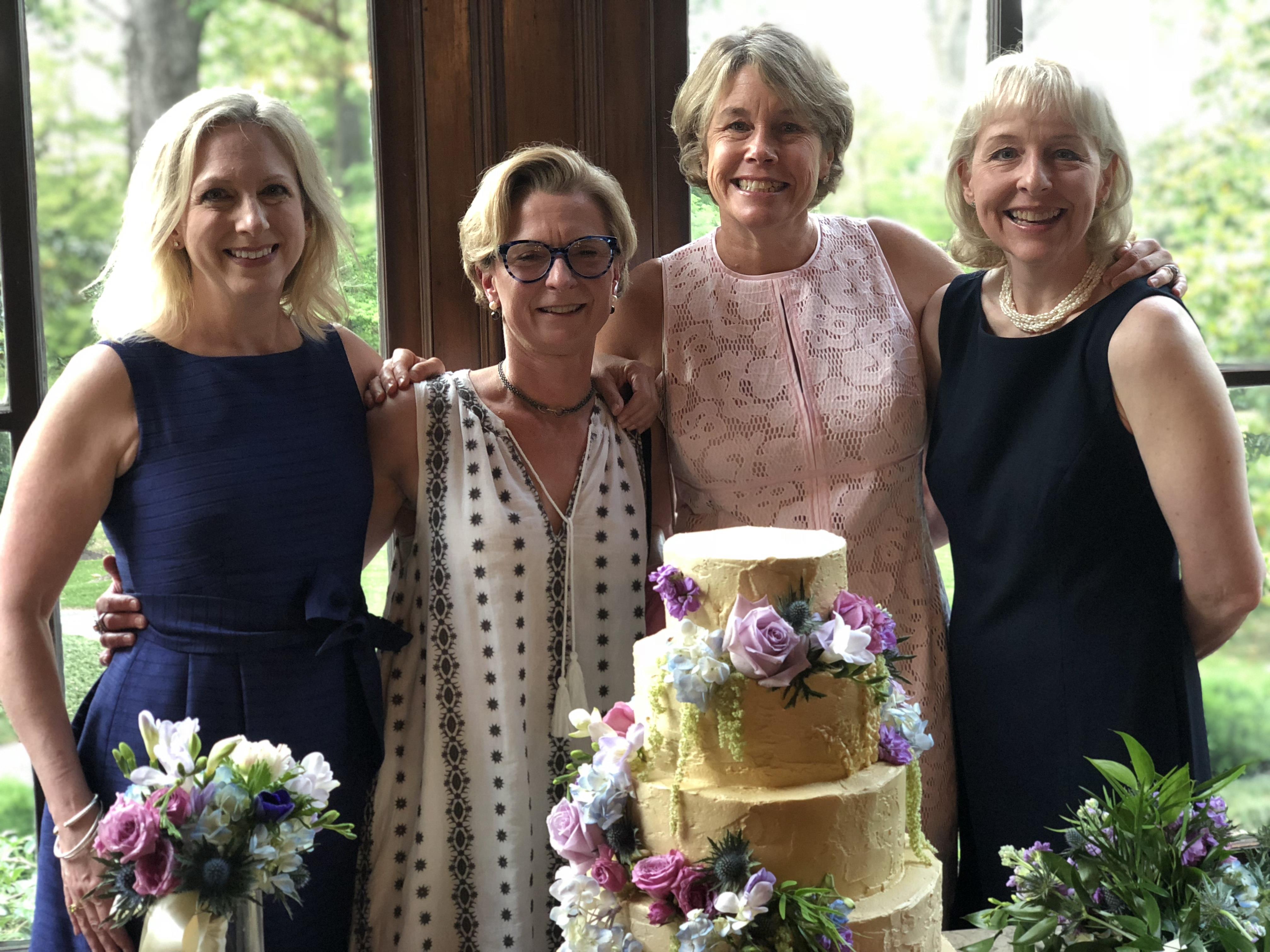 Friends gather indoors surrounded by flowers and a beautiful cake during a joyful celebration.