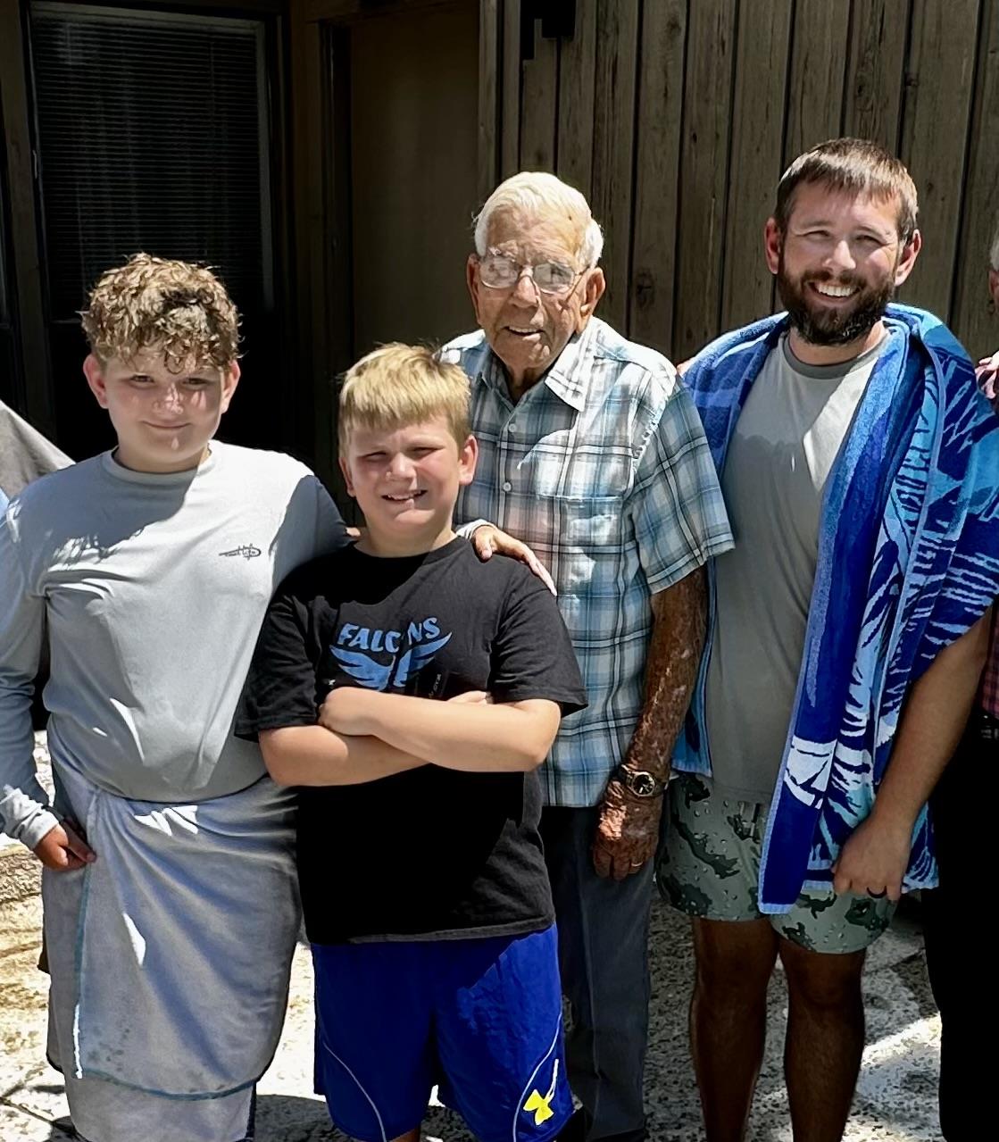 A group of three boys and their grandfather smile together outdoors on a sunny day.