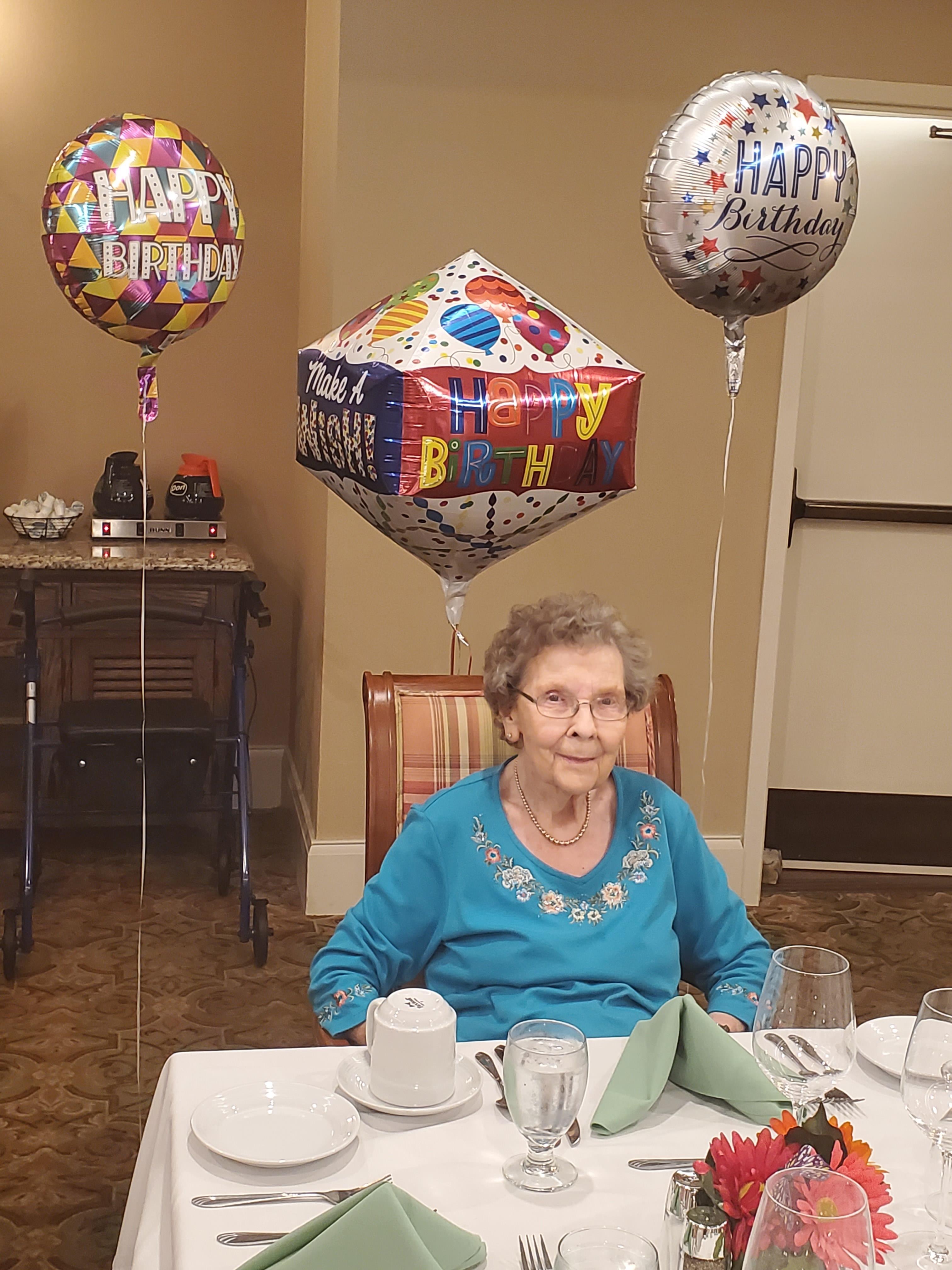 An elderly woman celebrates her birthday surrounded by colorful balloons in a cozy setting.