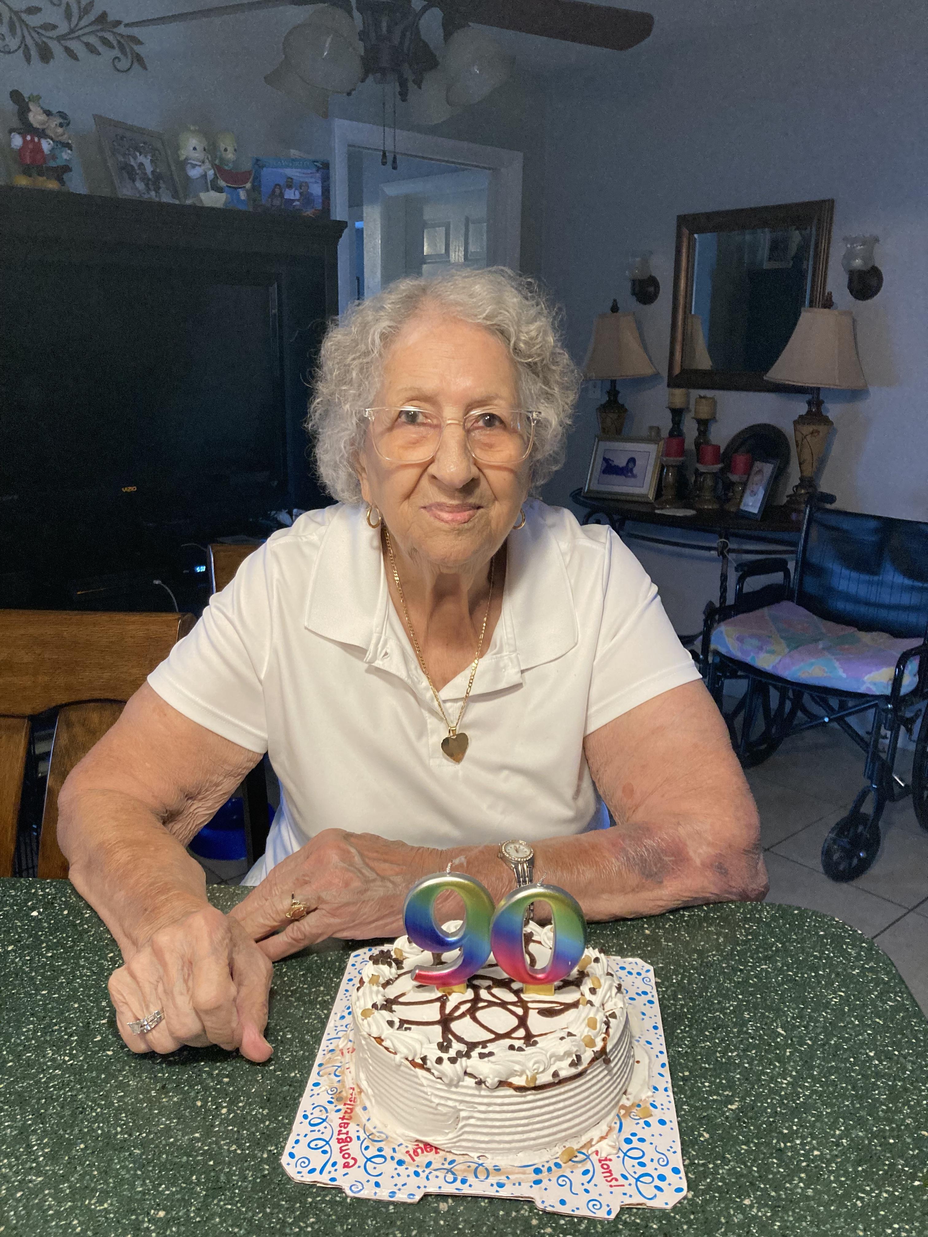 Elderly woman smiles proudly as she celebrates her birthday with a decorated cake in a cozy home.